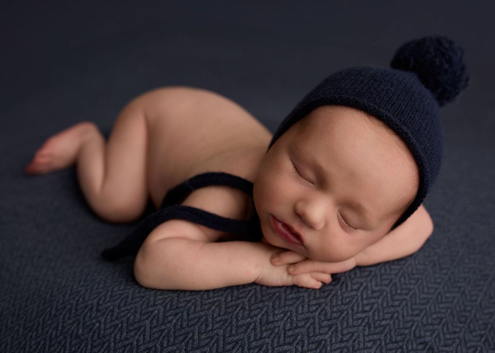 Sleeping newborn baby wearing a navy blue knit bonnet with pompom, posed on a blue textured blanket -- Sara Prue Photography Denver