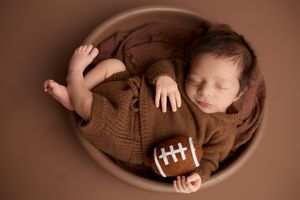 Newborn baby sleeping in a bowl wrapped in brown knit holding a mini football prop. -- Sara Prue Photography Denver