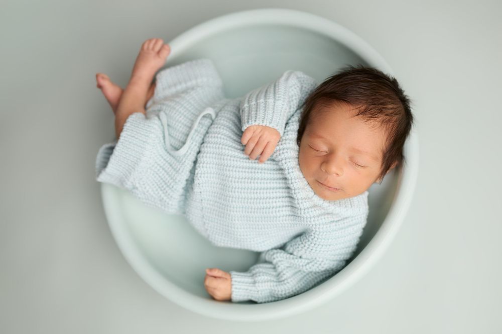 Newborn baby sleeping peacefully in a blue bowl, wearing a soft knit blue outfit, on a mint background. -- Sara Prue Photography Denver
