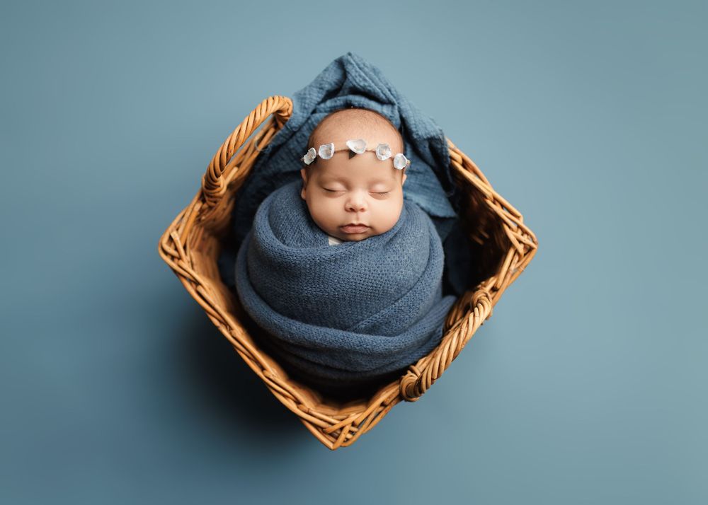 Sleeping newborn baby wrapped in blue swaddle with floral headband, nestled in a wicker basket -- -- Sara Prue Photography Denver