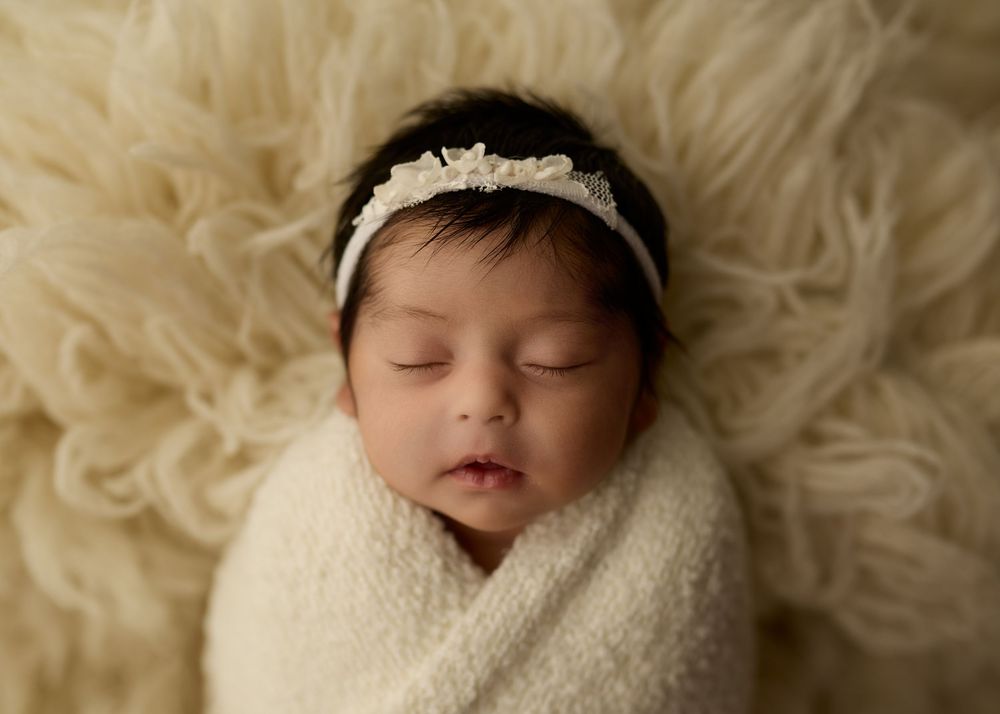 Sleeping newborn baby wrapped in cream knit swaddle with white floral headband on fluffy ivory fur backdrop.-- Sara Prue Photography Denver