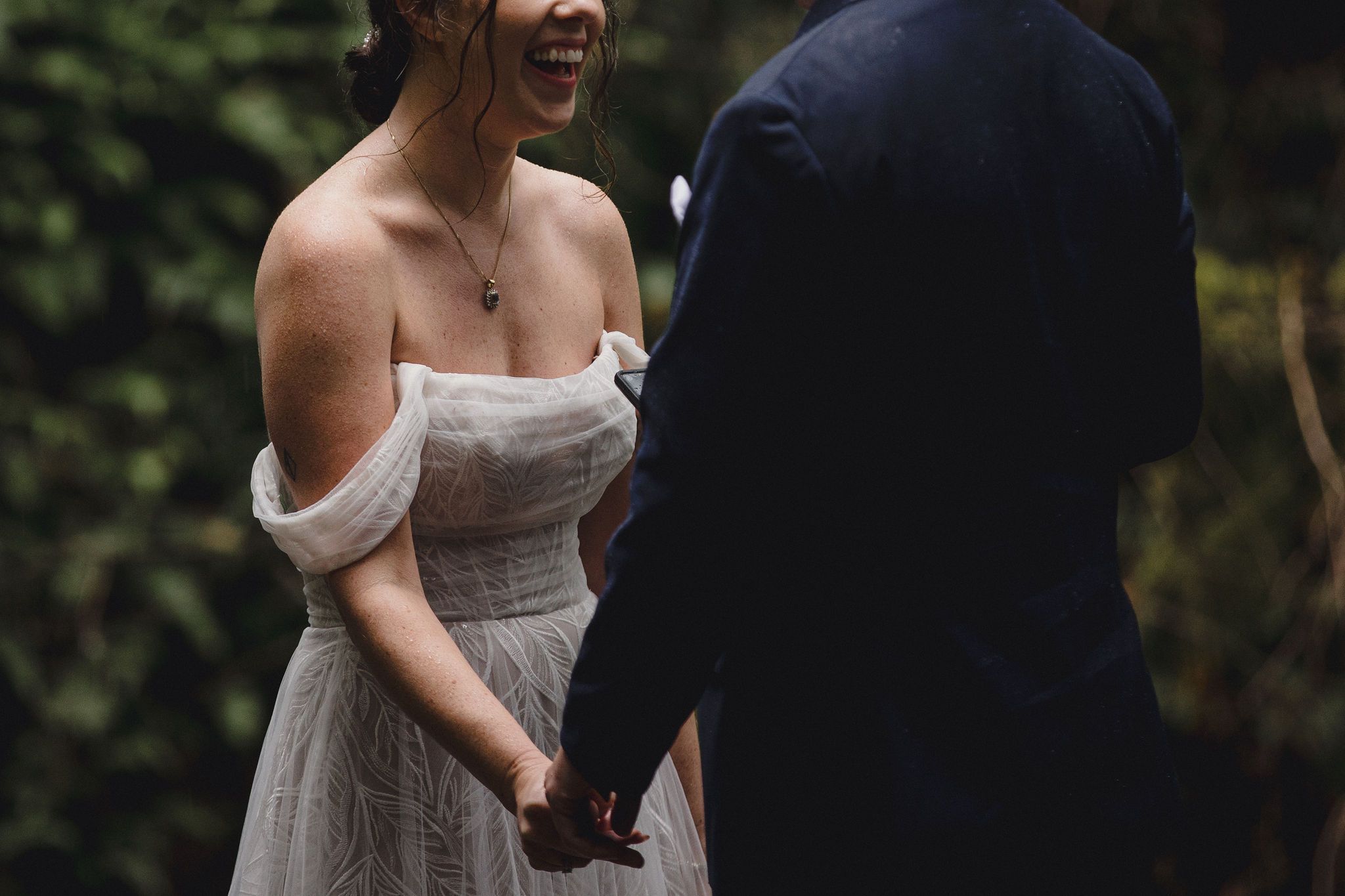 Bride in off-shoulder white gown laughing joyfully while holding hands with groom in navy suit during their Oregon elopement ceremony.