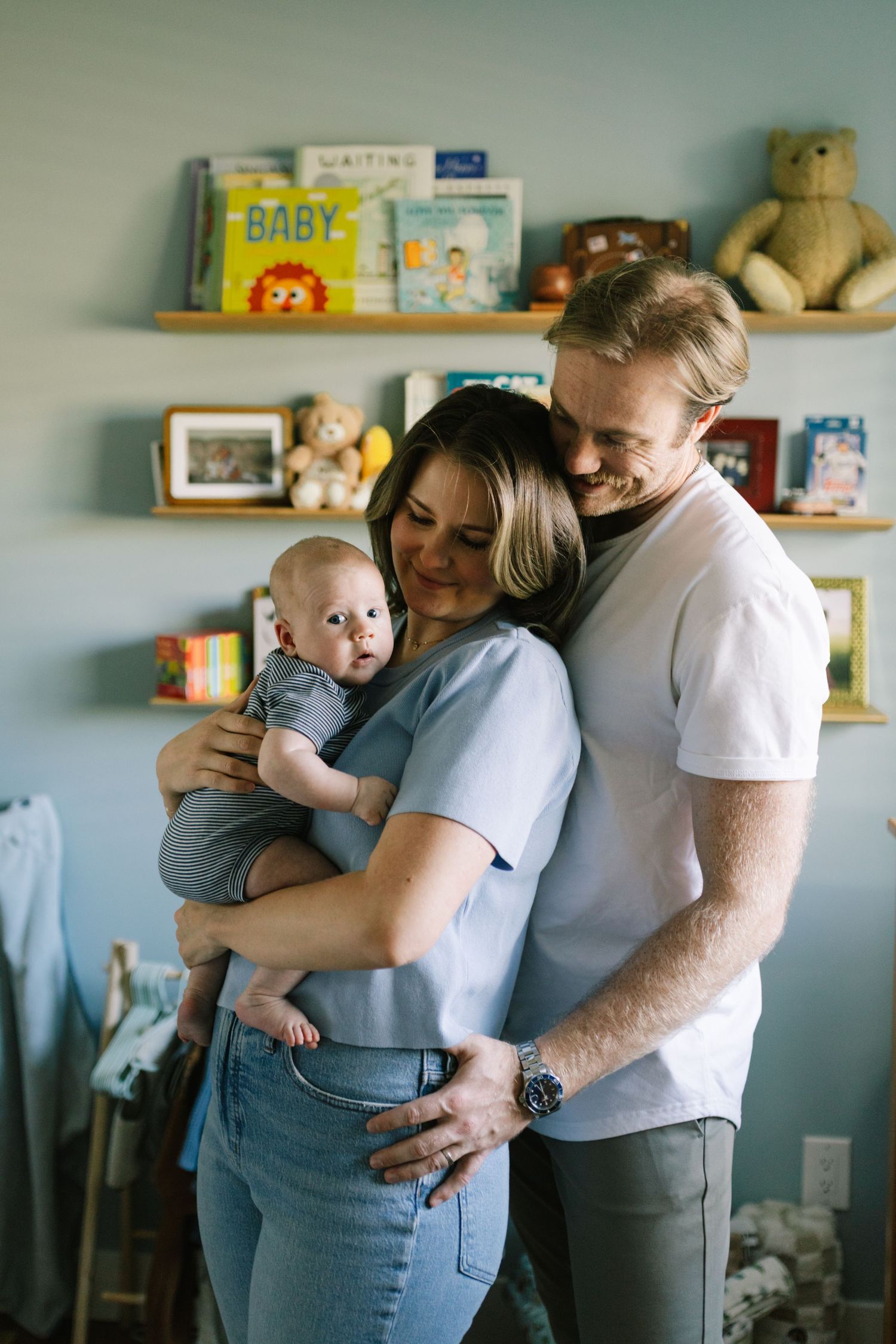Lifestyle newborn sessions in Utah. Parents smiling and holding their newborn baby in a cozy nursery with colorful books on wall shelves.