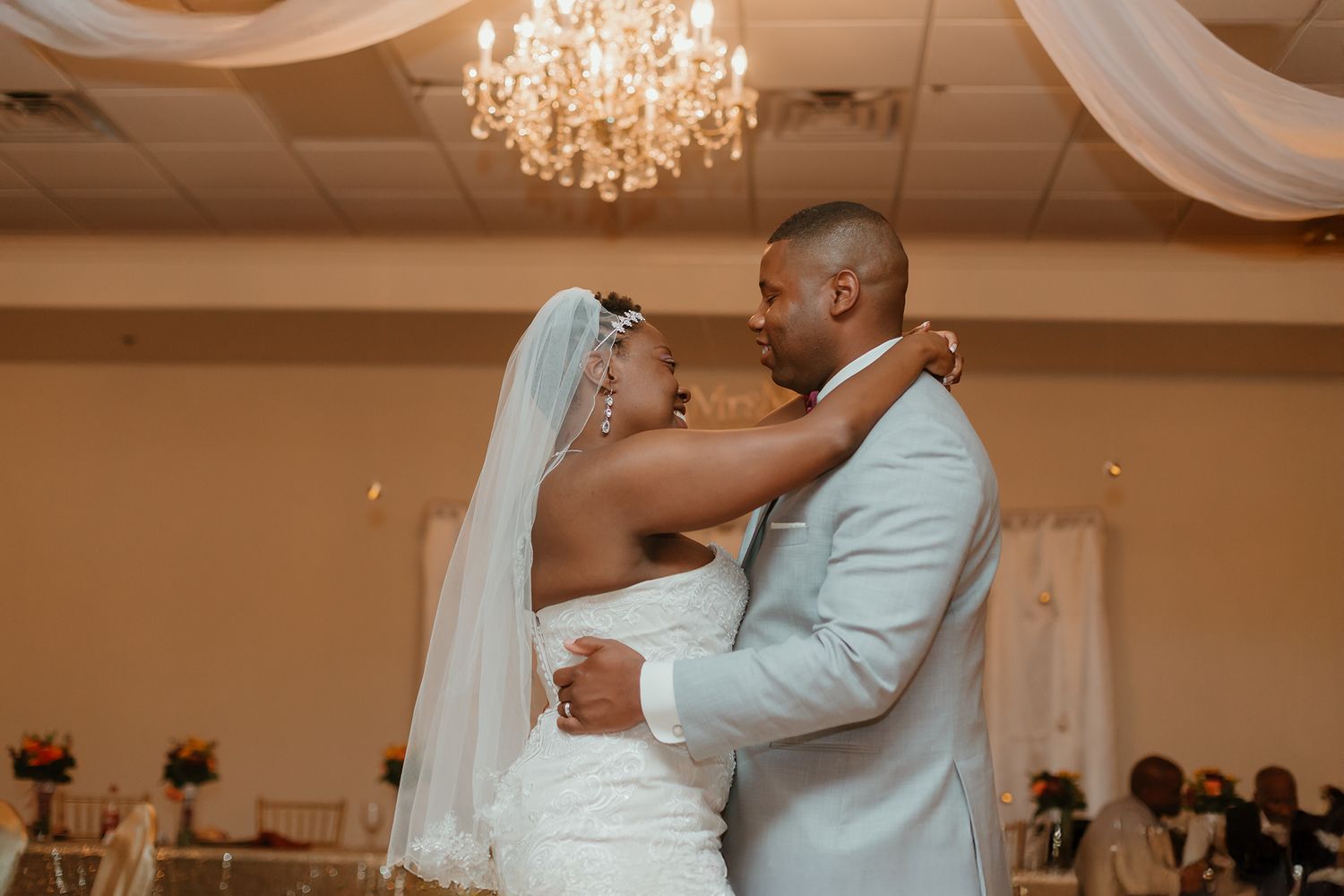 Newlywed couple sharing their first dance at wedding reception under elegant chandelier.