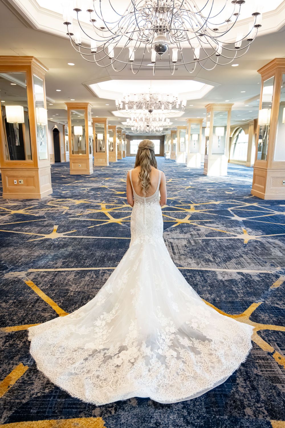 Bride in elegant lace mermaid wedding gown with long train stands in grand ballroom with crystal chandeliers.