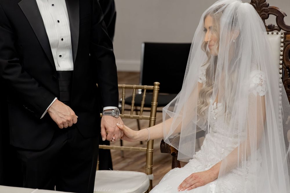 Bride in white gown and veil holding hands with groom in black tuxedo during wedding ceremony.