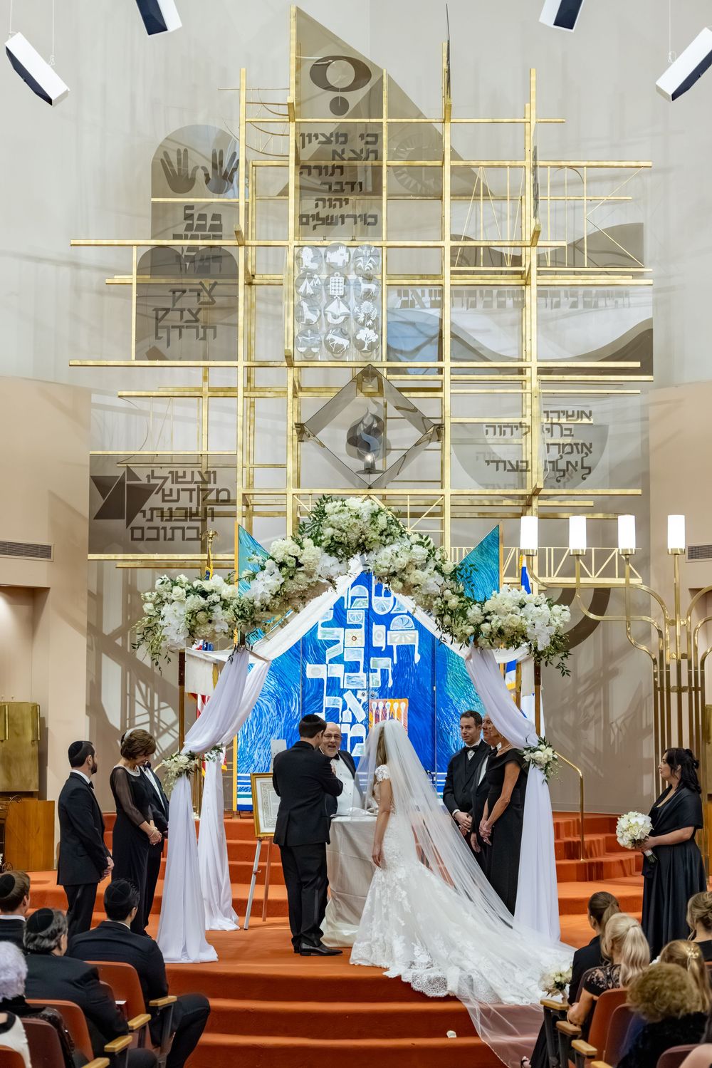 Jewish wedding ceremony under a floral chuppah in a synagogue with Hebrew inscriptions and gold architectural details.