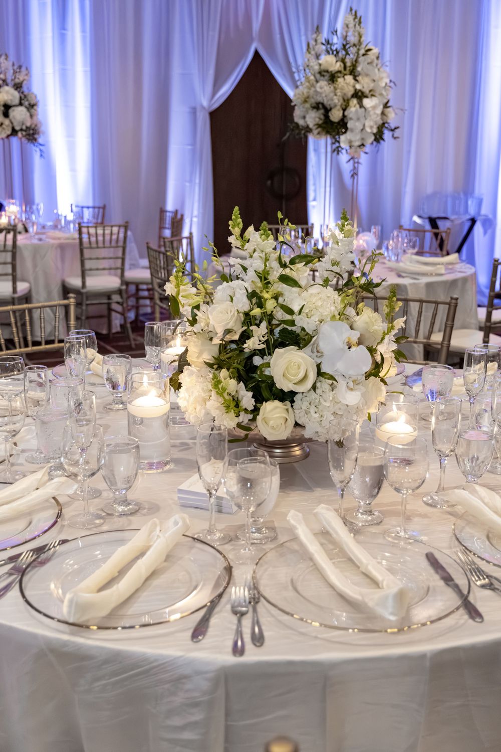 Elegant wedding reception table with white floral centerpiece, crystal glasses, silver chargers, and candles on white linen.