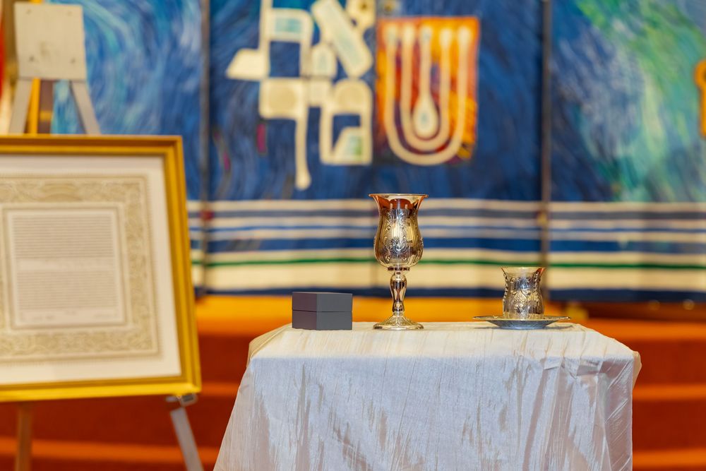 Jewish synagogue altar with silver Kiddush cup, small glass, and framed text on white draped table.
