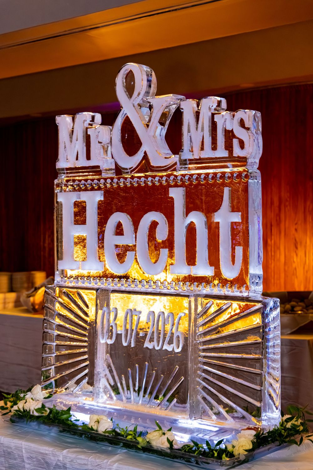 Illuminated ice sculpture reading Mr. & Mrs. Hecht dated 03-07-2025 at a wedding reception.