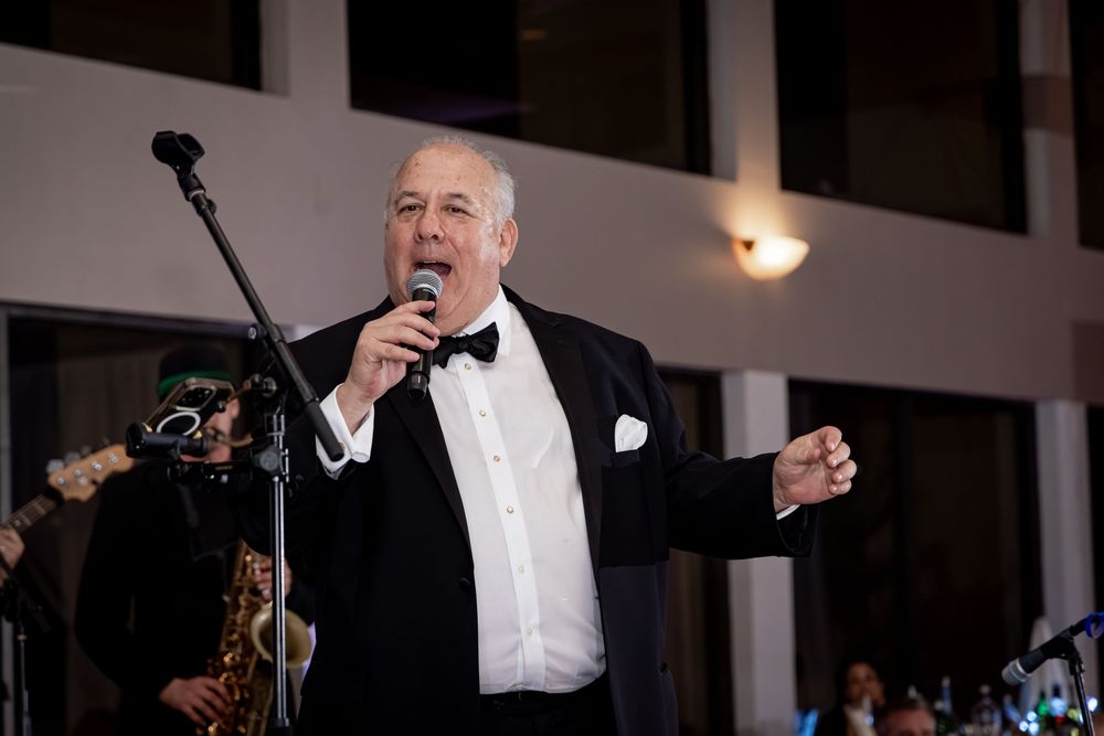 Elegant man in tuxedo singing into microphone at formal event with band musicians visible in background.