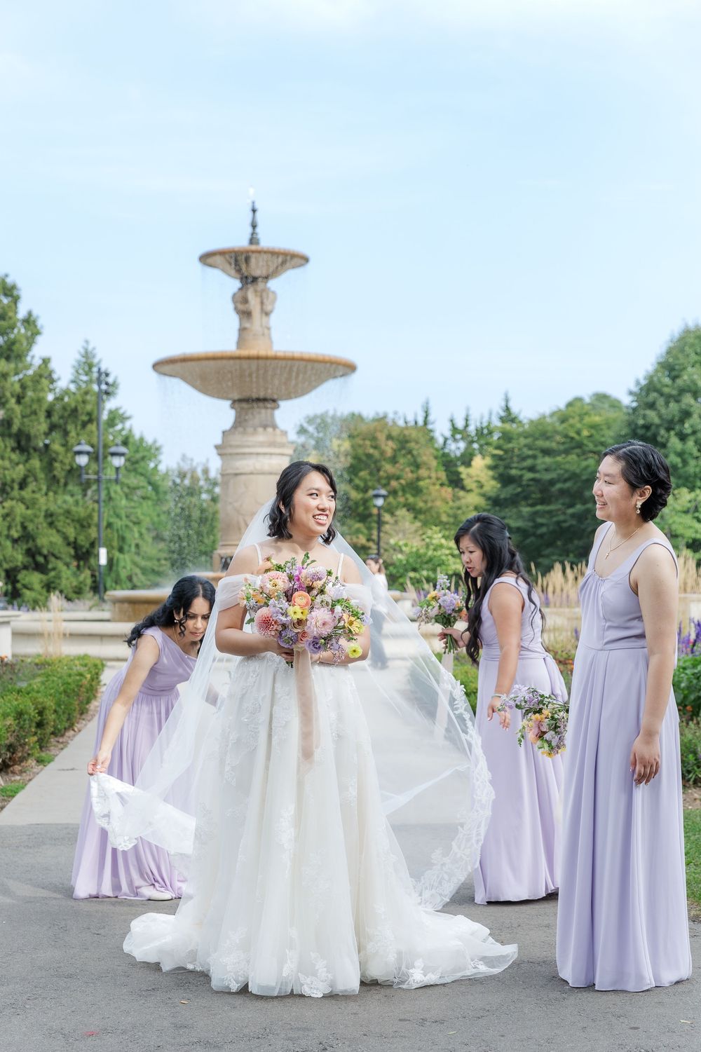 Candid wedding photo of bridesmaids adjusting the brides dress in front of the fountain in gage park in hamilton, ontario.