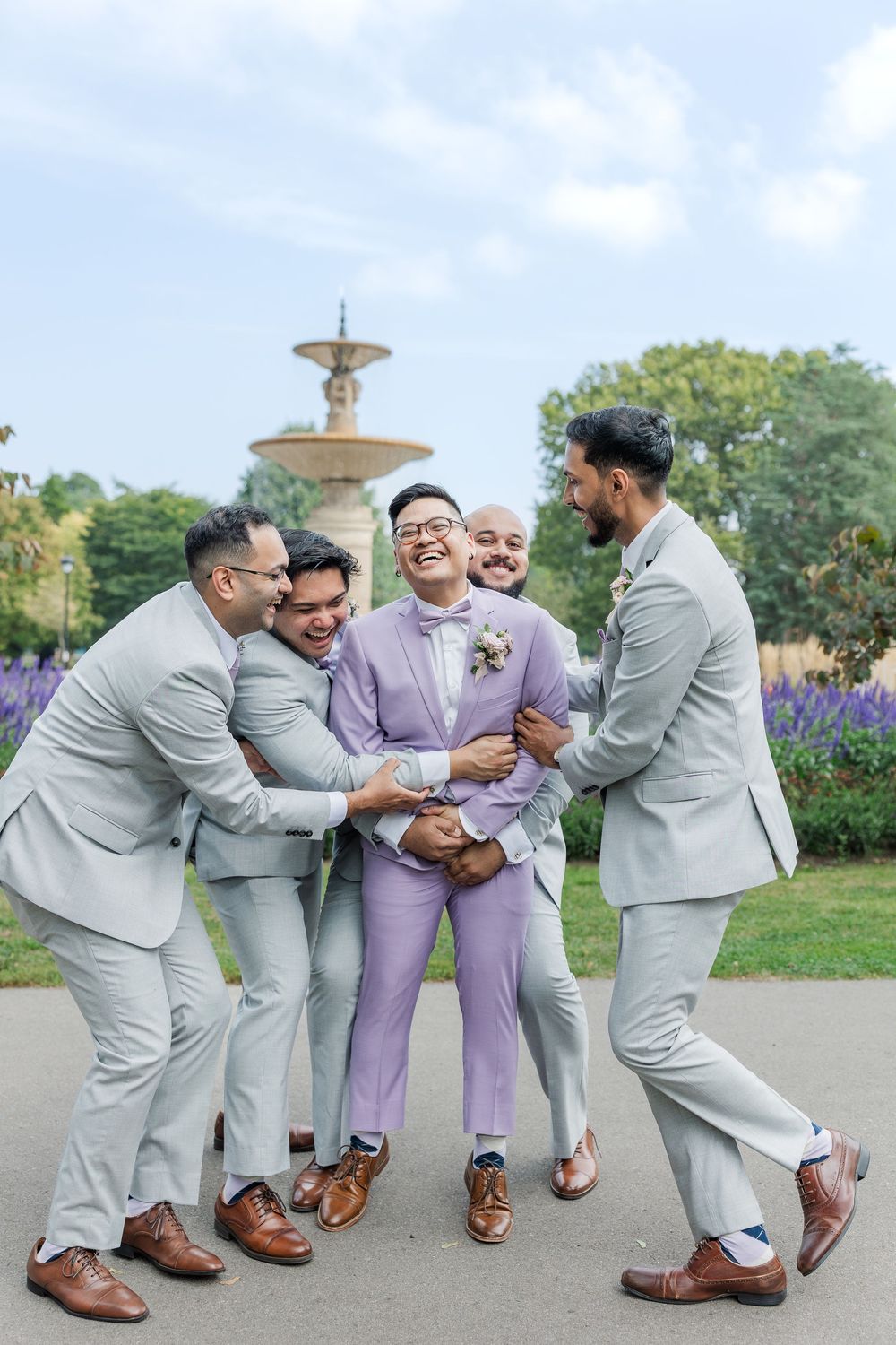 Candid wedding photo of the groomsmen hugging the groom in front of the fountain in gage park during their hamilton, ontario wedding.