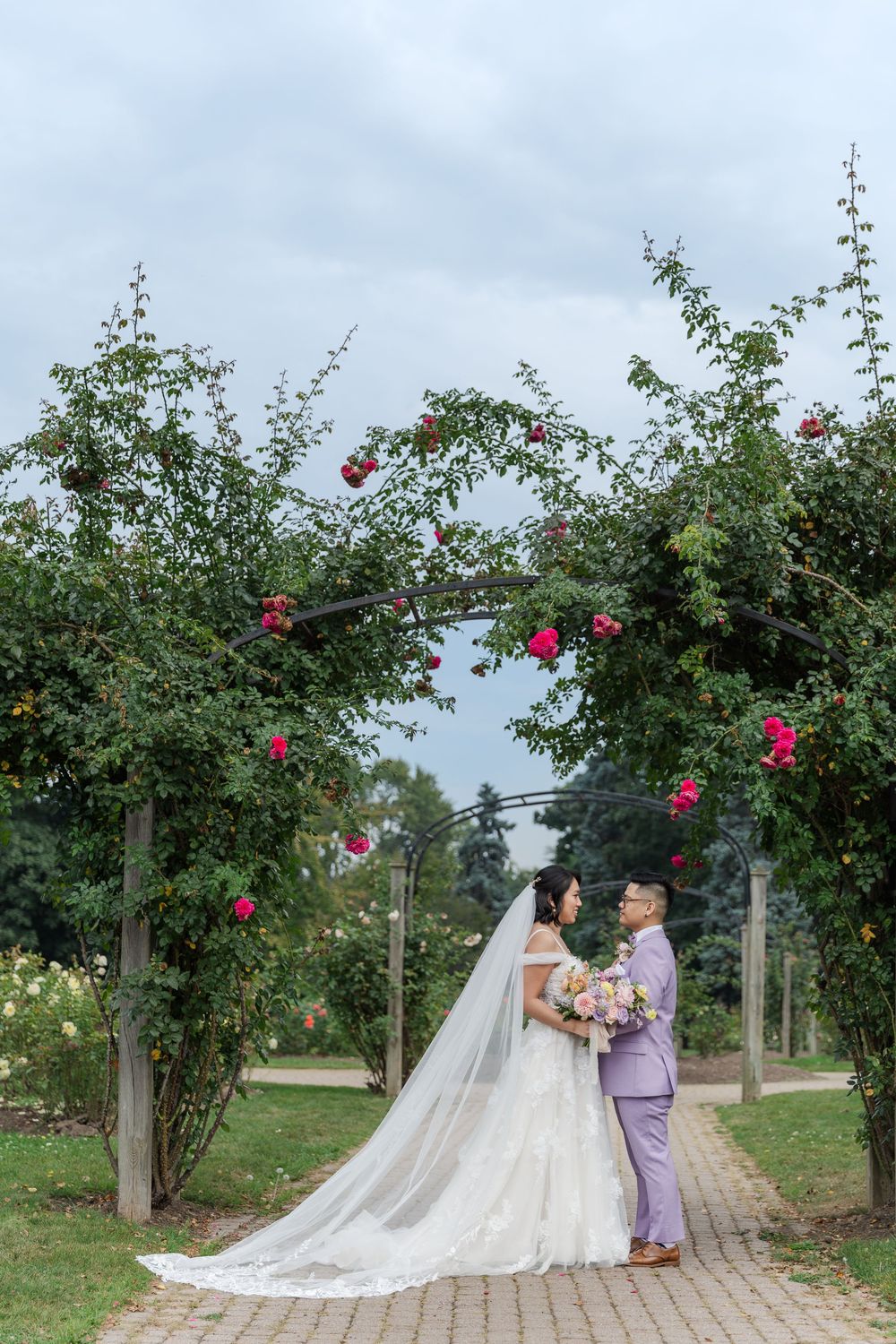 Calm wedding photo of bride and groom standing together below an arch of red roses in the gardens at gage park in hamilton, ontario.