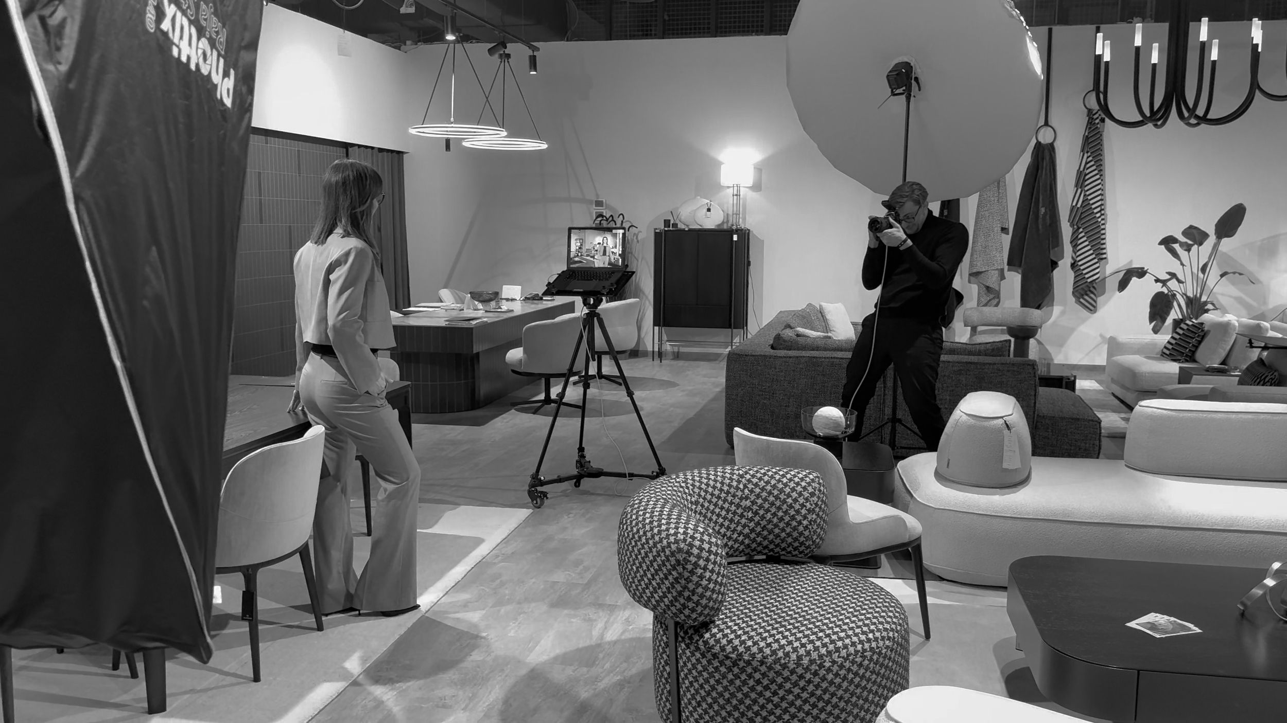 Czarno-białe zdjęcie biznesowej sesji fotograficznej w salonie meblowym z modelami, sprzętem oświetleniowym oraz krzesłami i sofami.
