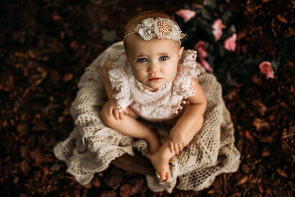 Overhead view of a baby wearing a white lace dress and floral headband sitting on a textured blanket.
