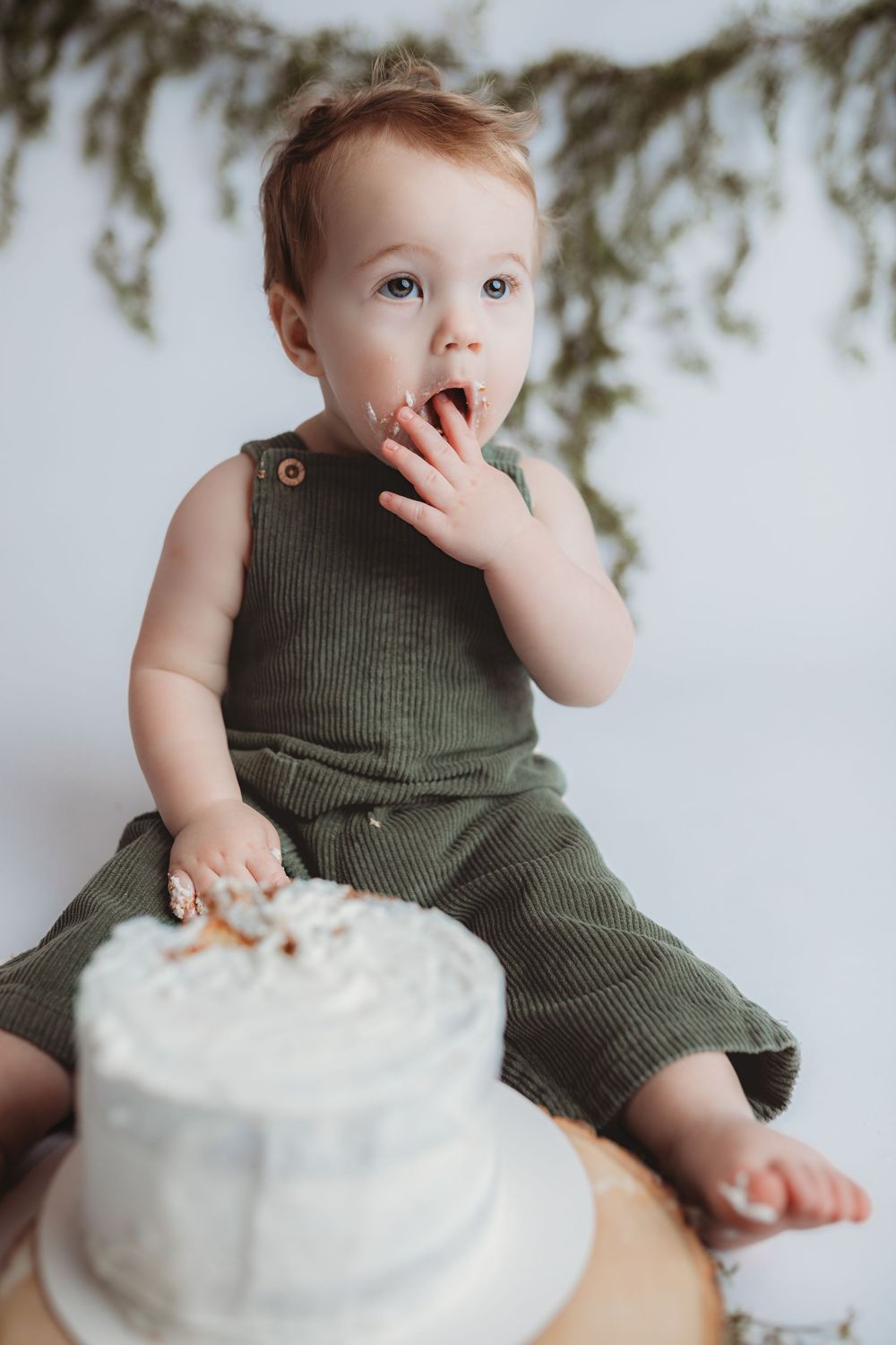Baby in olive overalls reaches toward white cake while sitting against wall with dried botanicals.