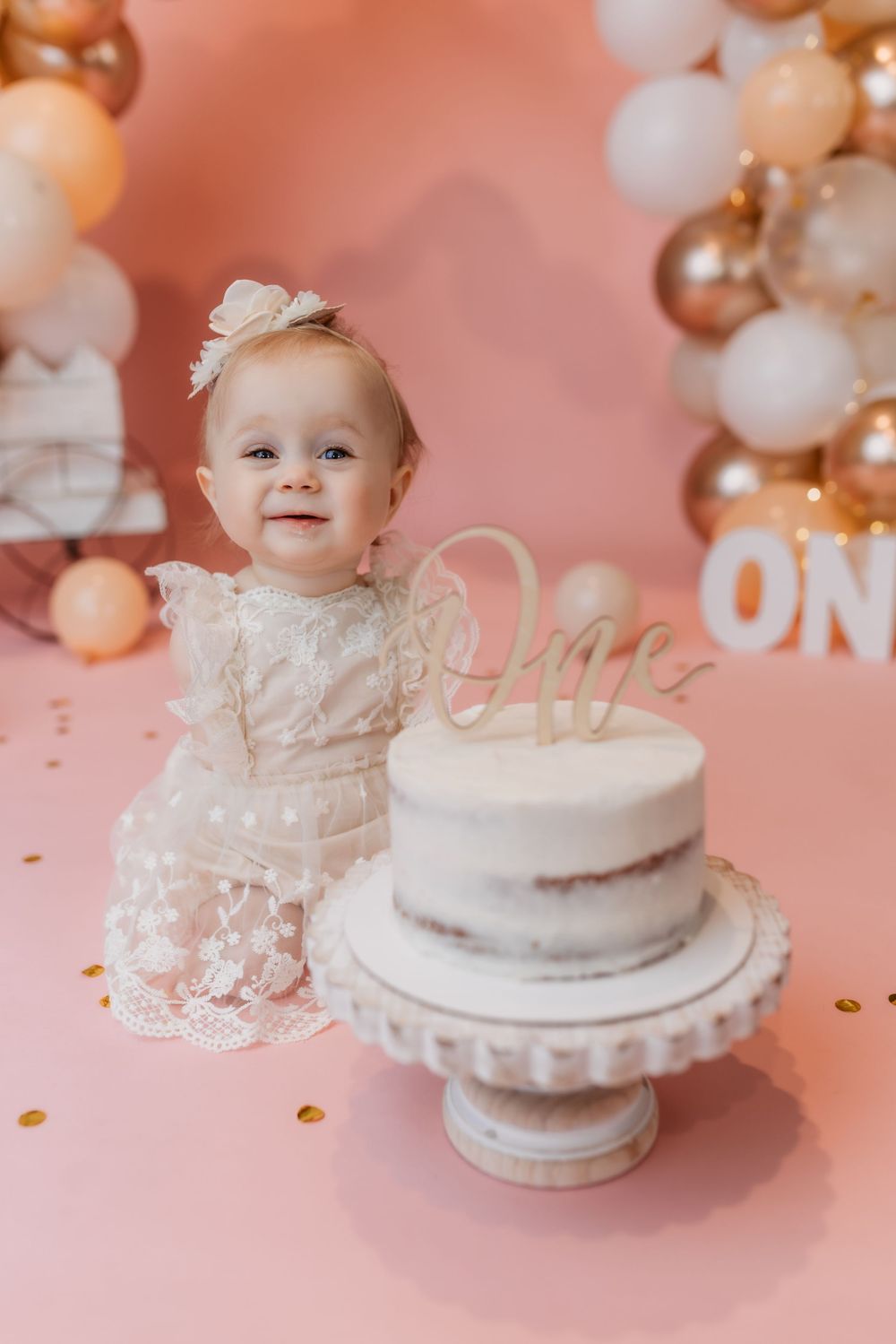 Baby girl in white lace dress with bow headband beside first birthday cake on pink backdrop.