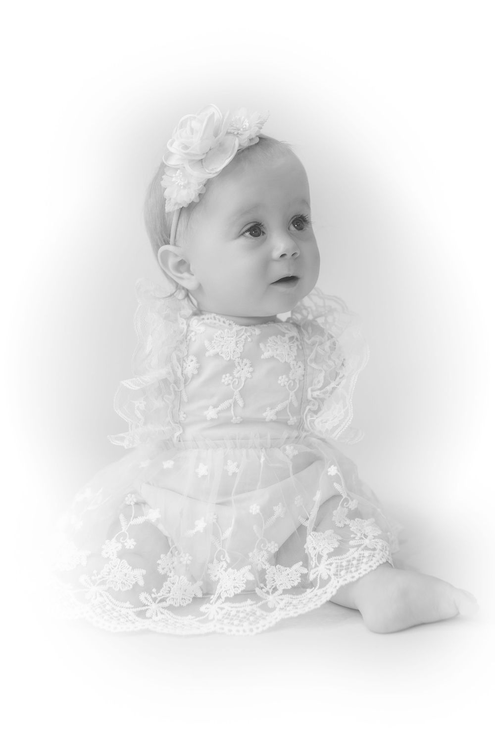 Black and white portrait of a baby girl in a lace dress and floral headband, sitting gracefully.