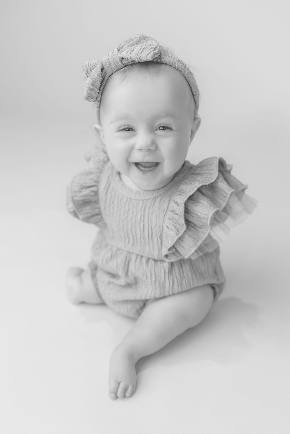 Black and white photo of a laughing baby girl in a ruffled romper and bow headband sitting up.