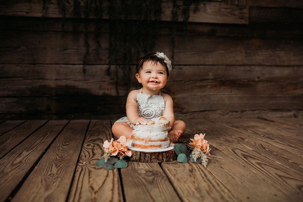 Baby in white dress sitting on rustic wooden floor for first birthday cake smash photoshoot.