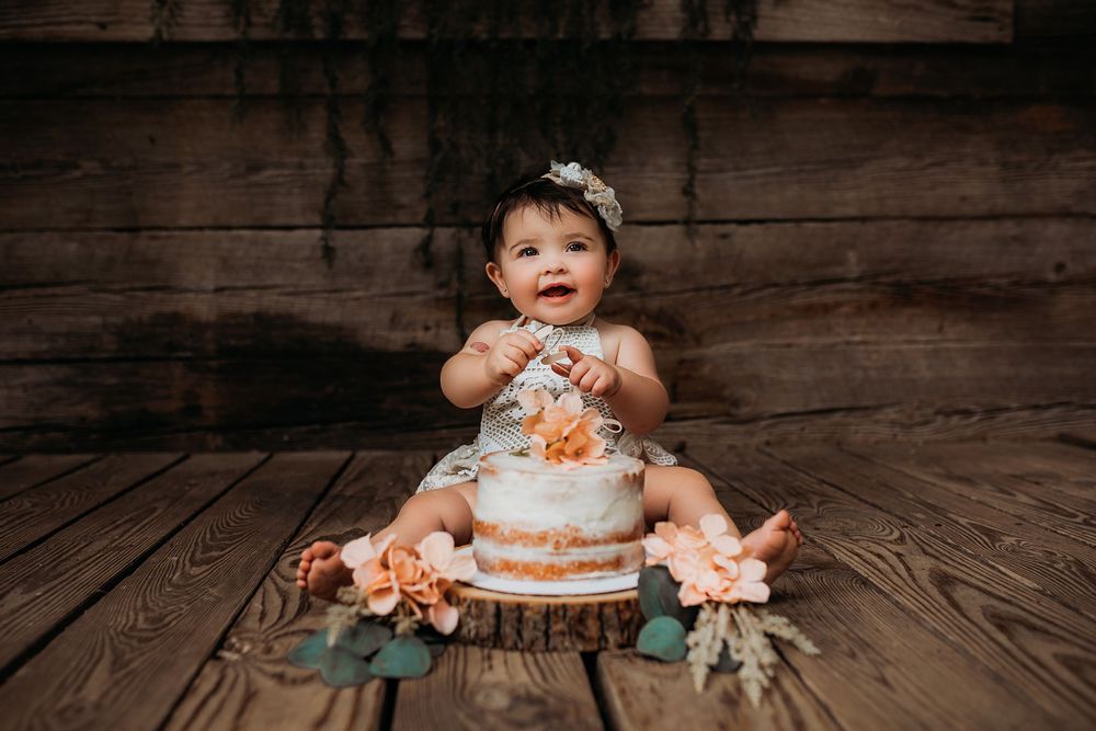 A baby in a blue dress sits on rustic wooden floorboards with vintage decor and soft lighting.