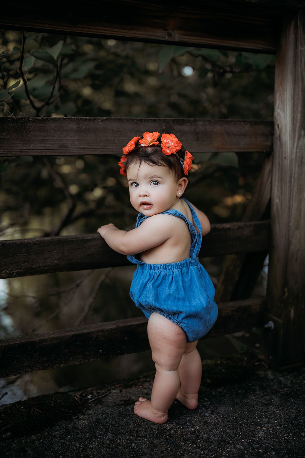 A toddler wearing a blue romper and orange flower crown looks back while standing by a wooden fence.