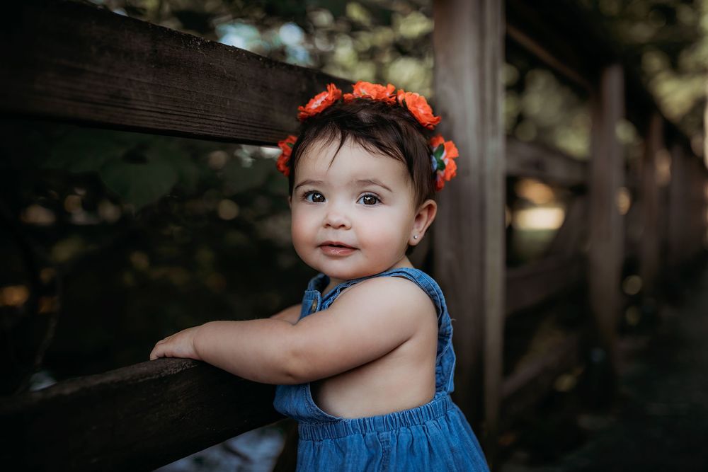 A young child in blue overalls and orange floral headband smiles while standing by a rustic wooden fence.