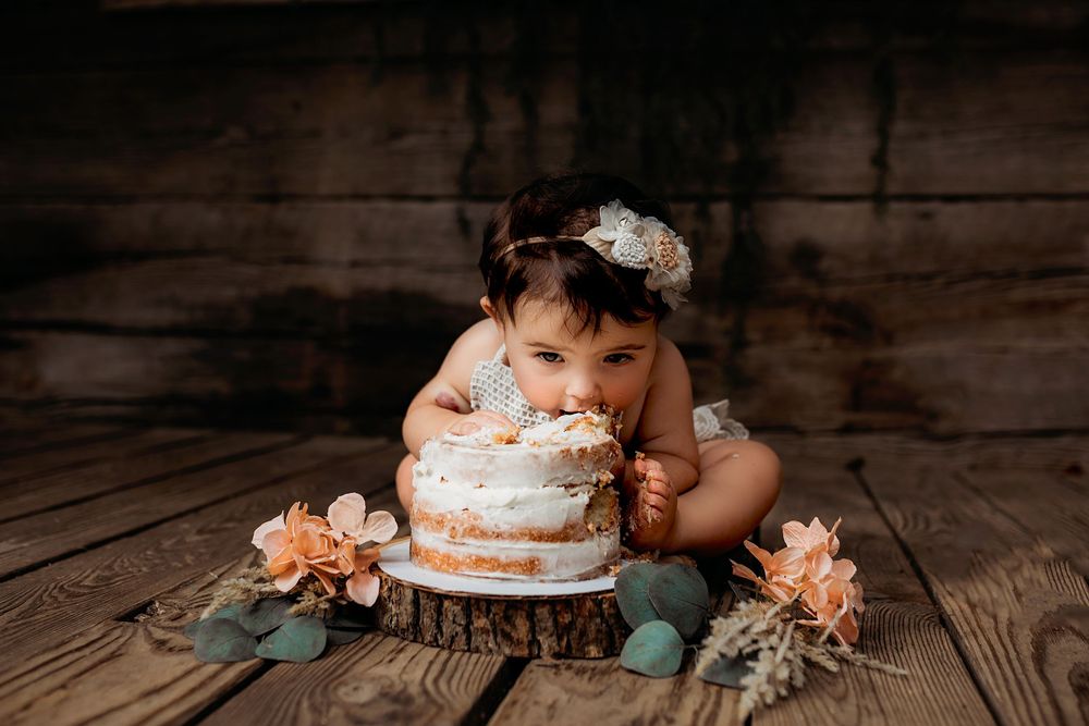 First birthday cake with peach flowers sits on rustic wooden floor during milestone photoshoot.