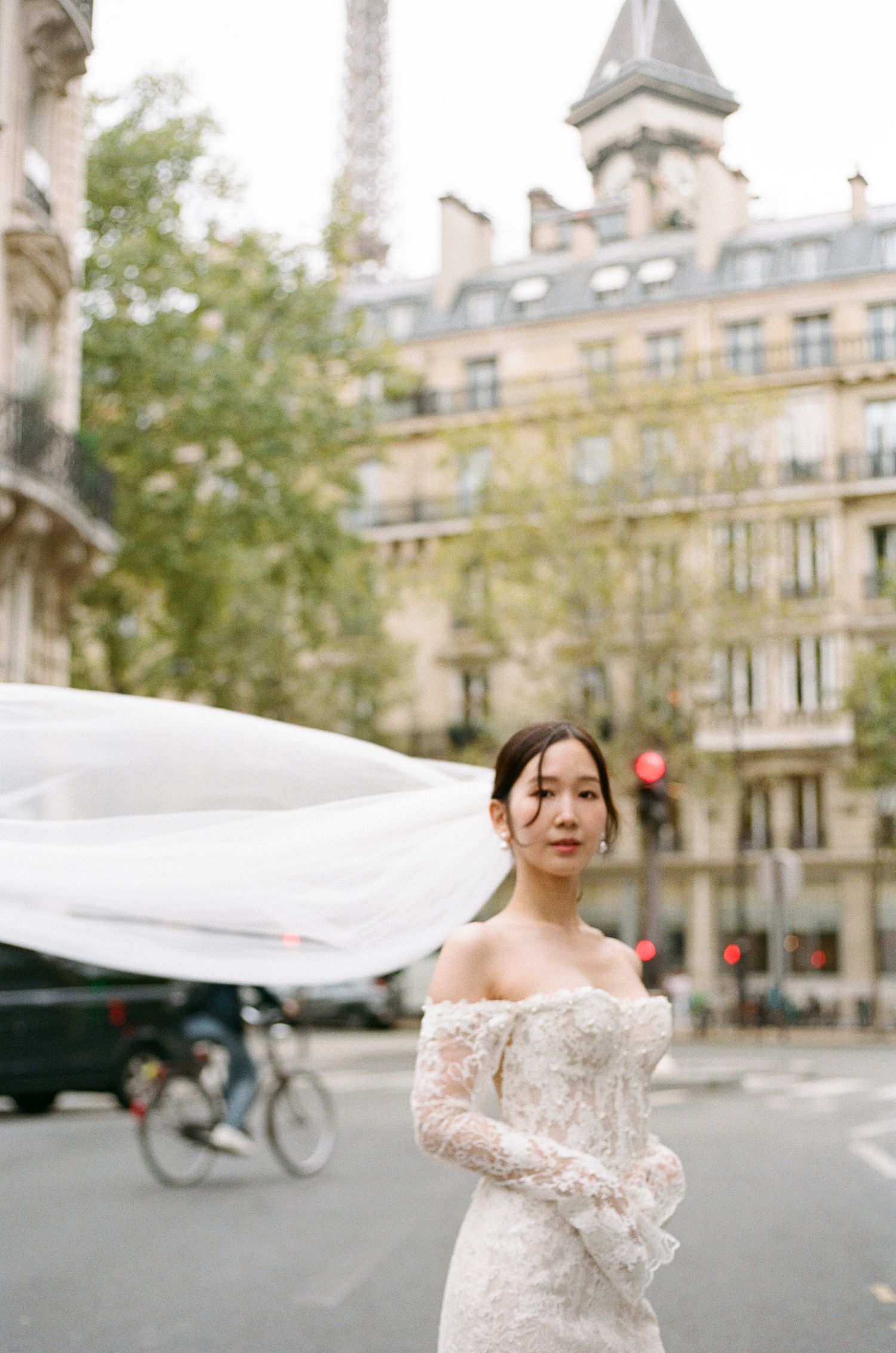 Bride in lace off-shoulder gown with flowing veil on Paris street, Eiffel Tower visible in background. Paris film photography, pre-wedding