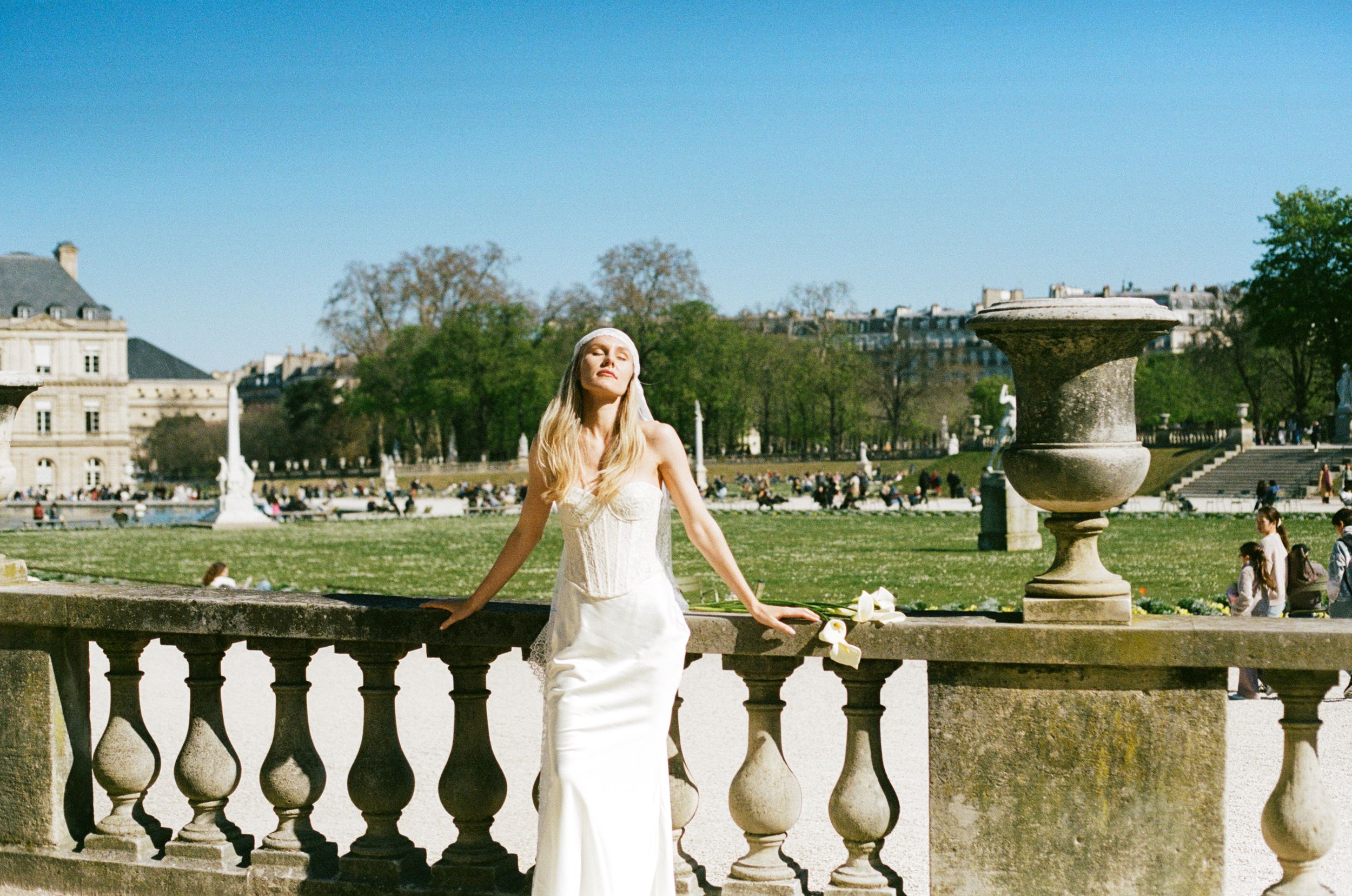 Bride in white gown posing at Luxembourg Gardens balustrade, Paris, on a sunny day with lush greenery.