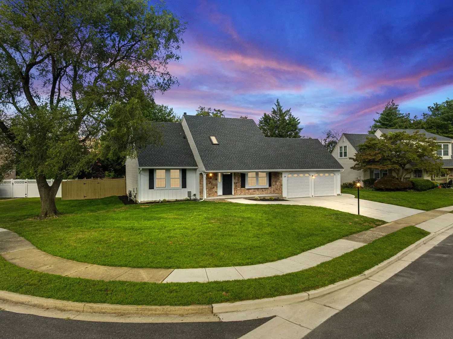 White cape cod home with attached garage, lush green lawn, mature trees, and a stunning sunset sky in a quiet neighborhood.
