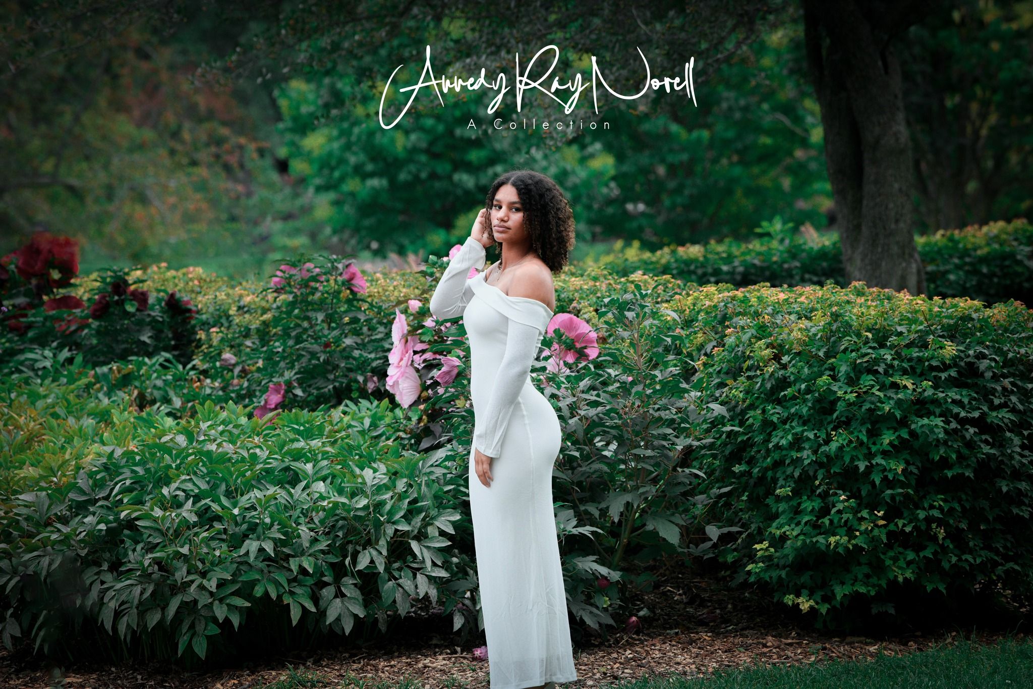 A woman in a white off-shoulder gown poses gracefully among lush green gardens with pink blooms.
