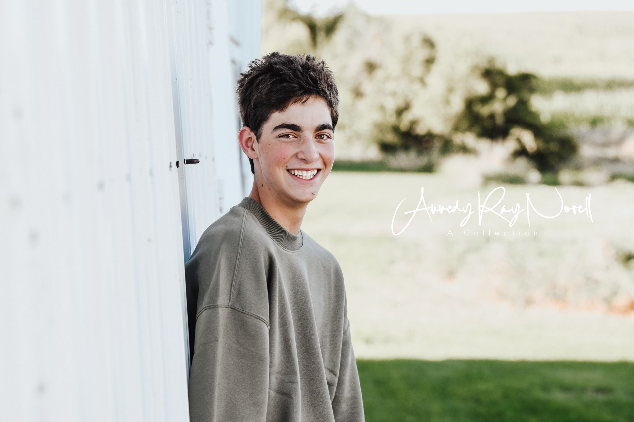 Smiling teenage boy in olive sweatshirt leaning against white barn wall outdoors.
