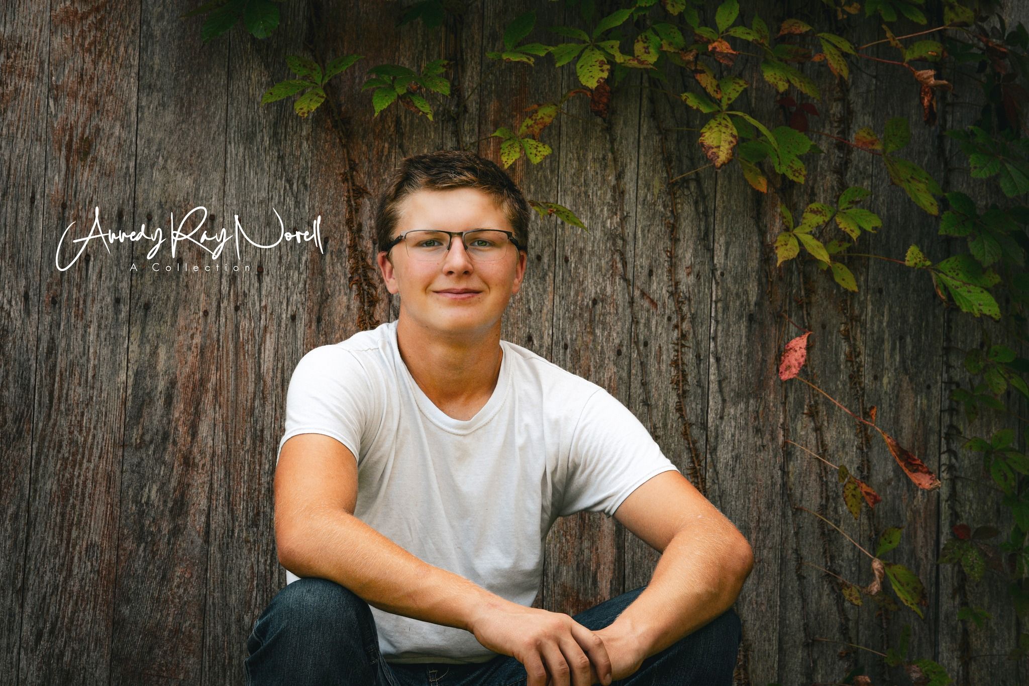 Young man in glasses and white t-shirt smiling while sitting against a large tree with ivy leaves.