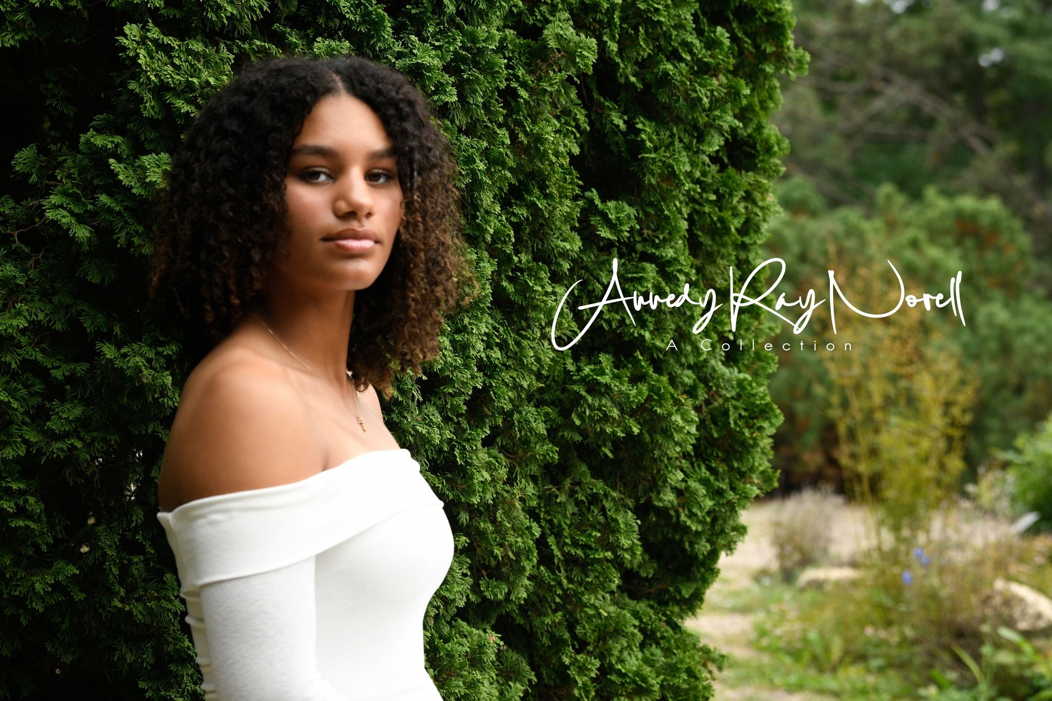 Young woman with curly hair in white off-shoulder top poses gracefully against lush green ivy wall outdoors.