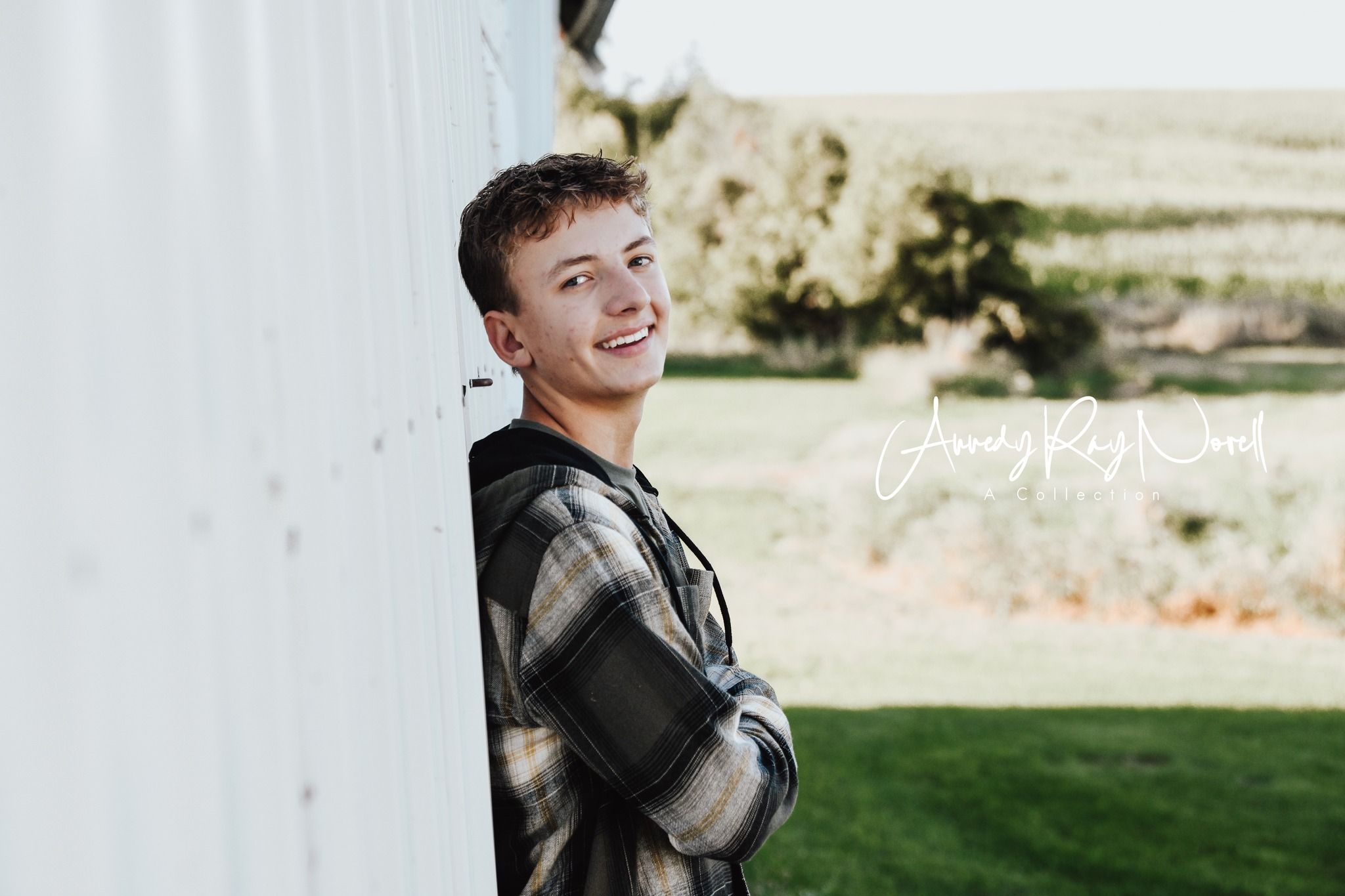 Smiling teenage boy in plaid shirt leaning against white barn wall with green countryside background.