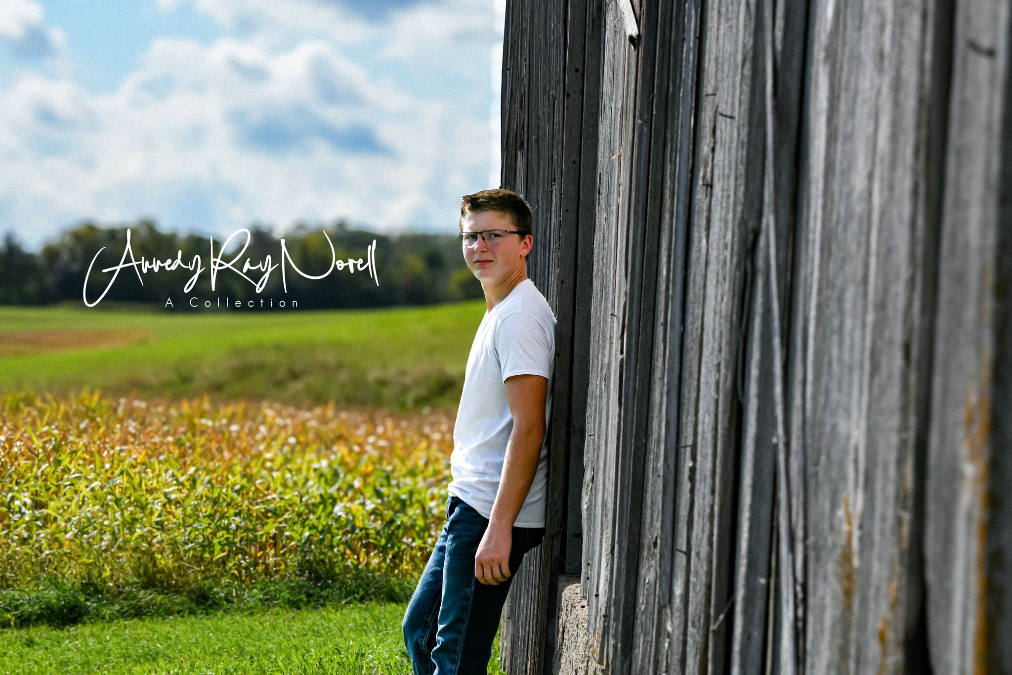 Teen boy leaning against rustic barn wall in rural field, outdoor senior portrait by Amanda Ray Wood.