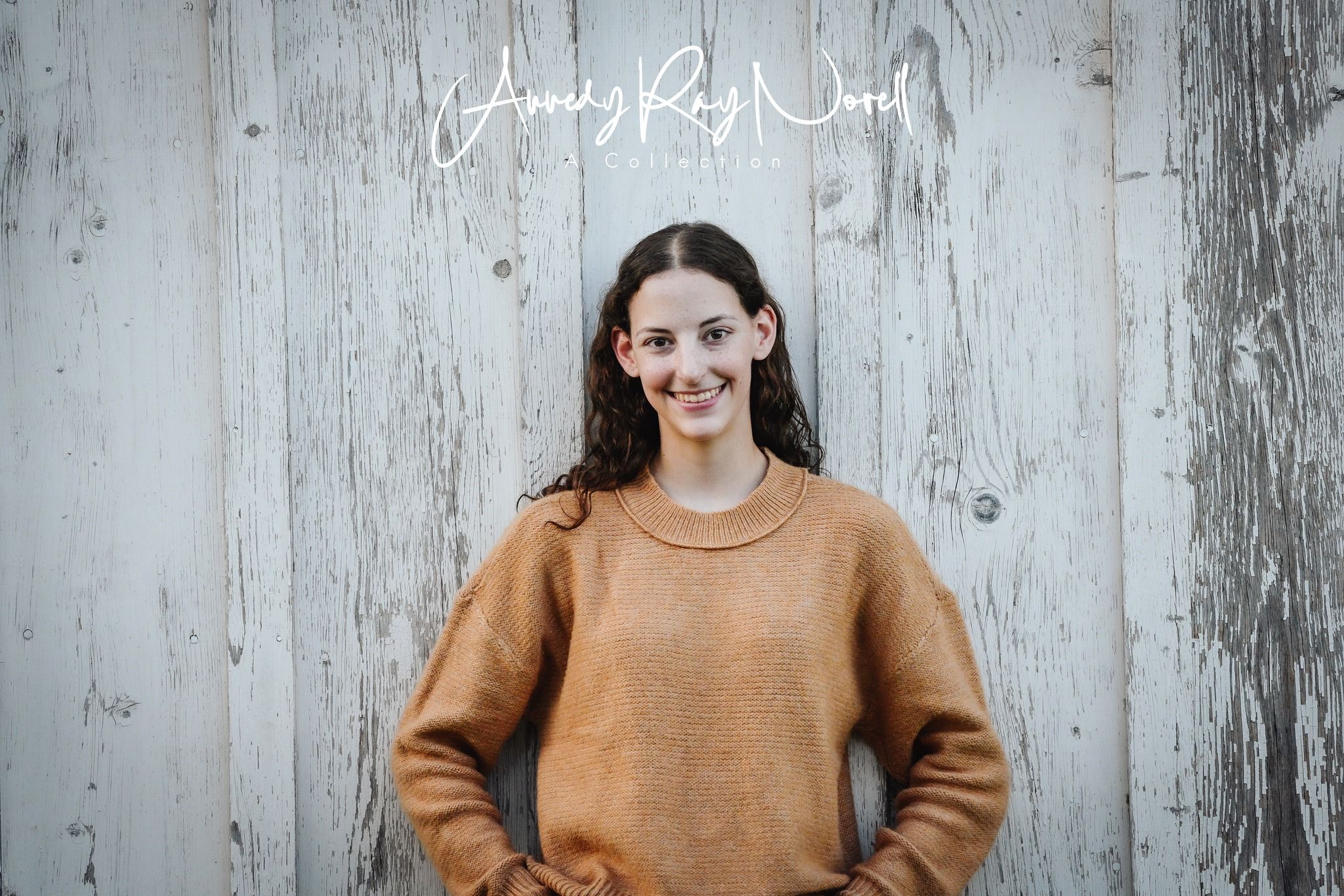 Smiling young woman in a camel sweater poses against a rustic white wooden background.