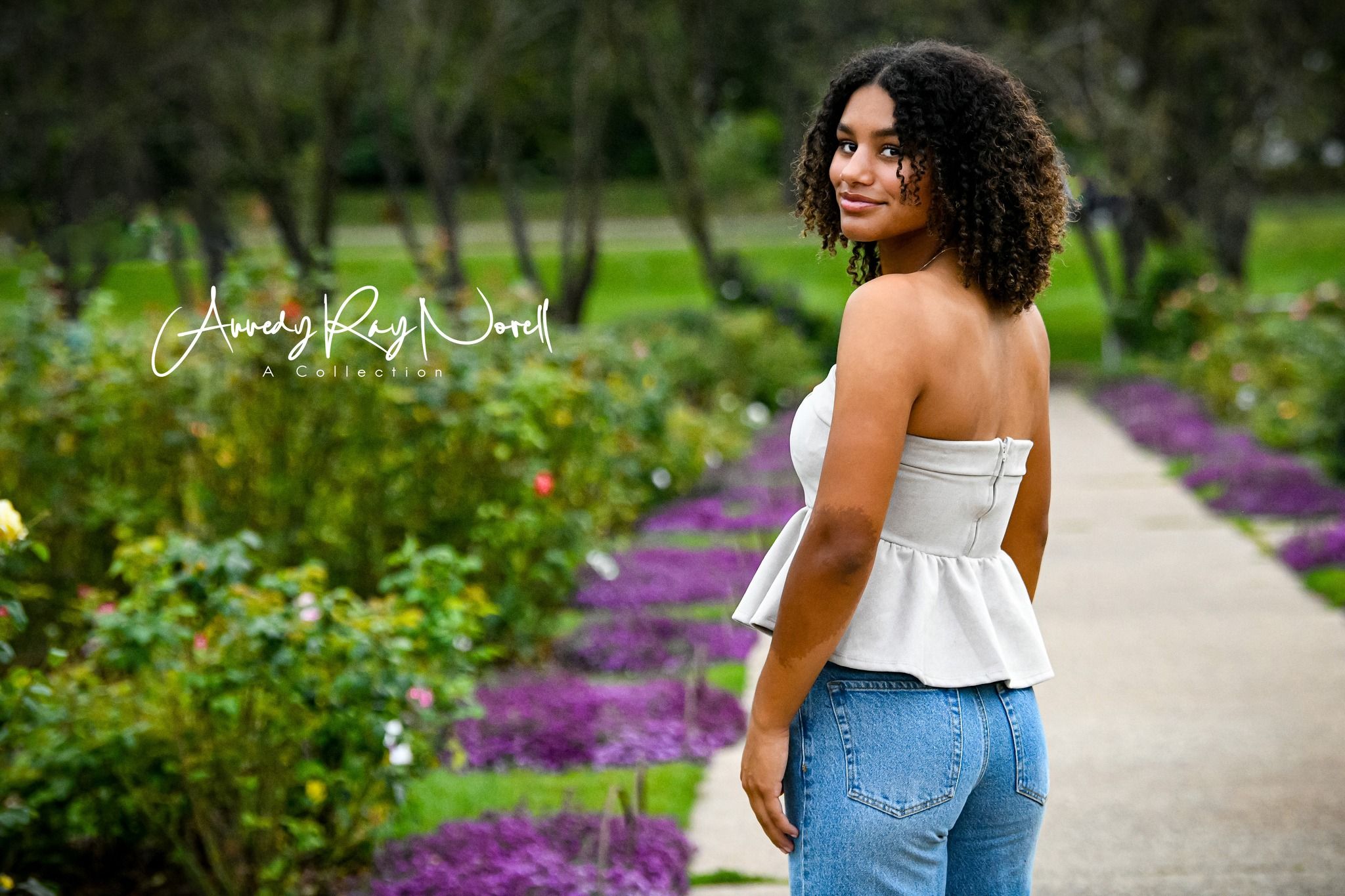 Young woman in white strapless top and jeans poses on garden path surrounded by colorful flowers.