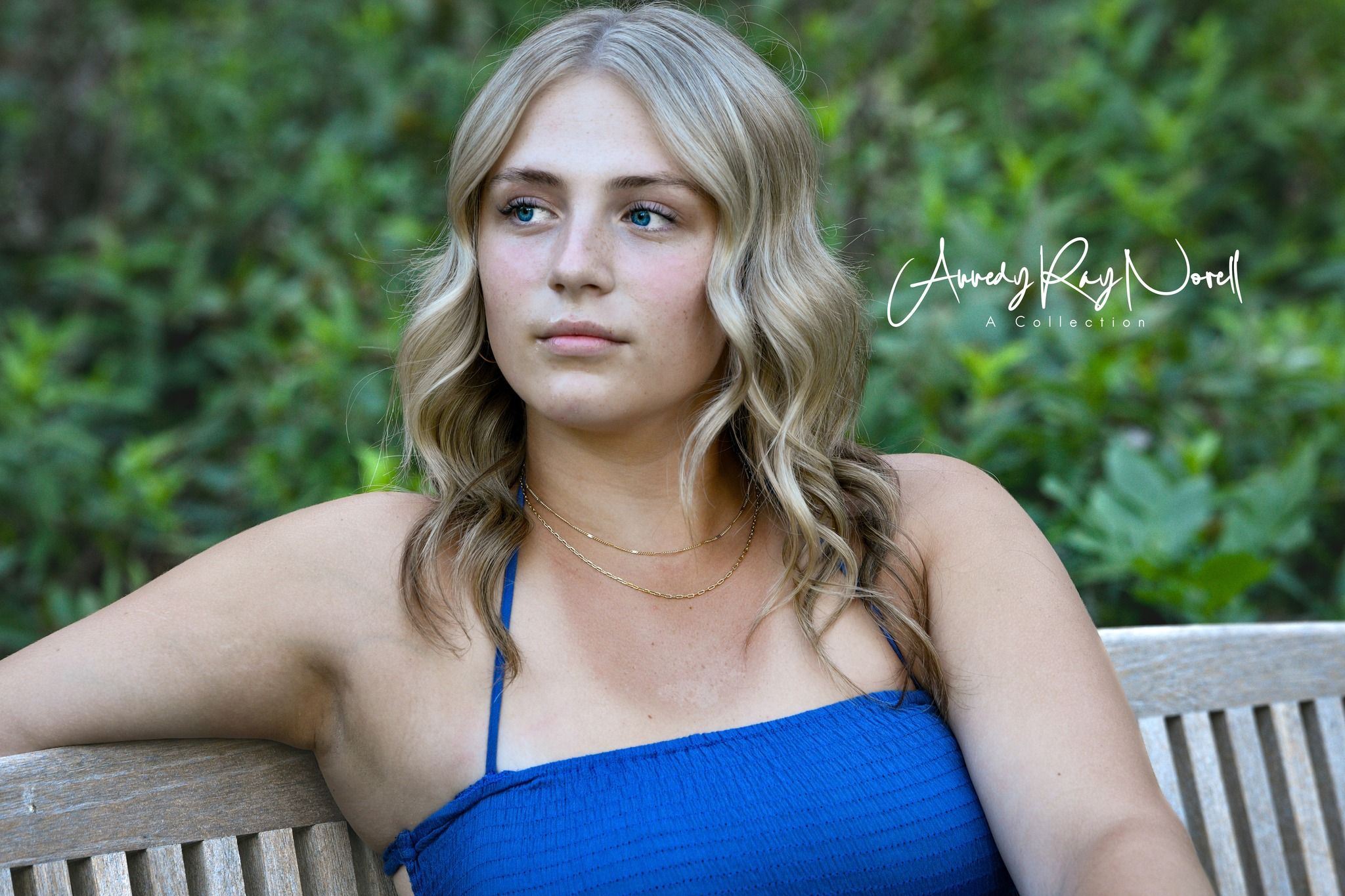 Young blonde woman in blue top sitting on wooden bench with lush green foliage background.