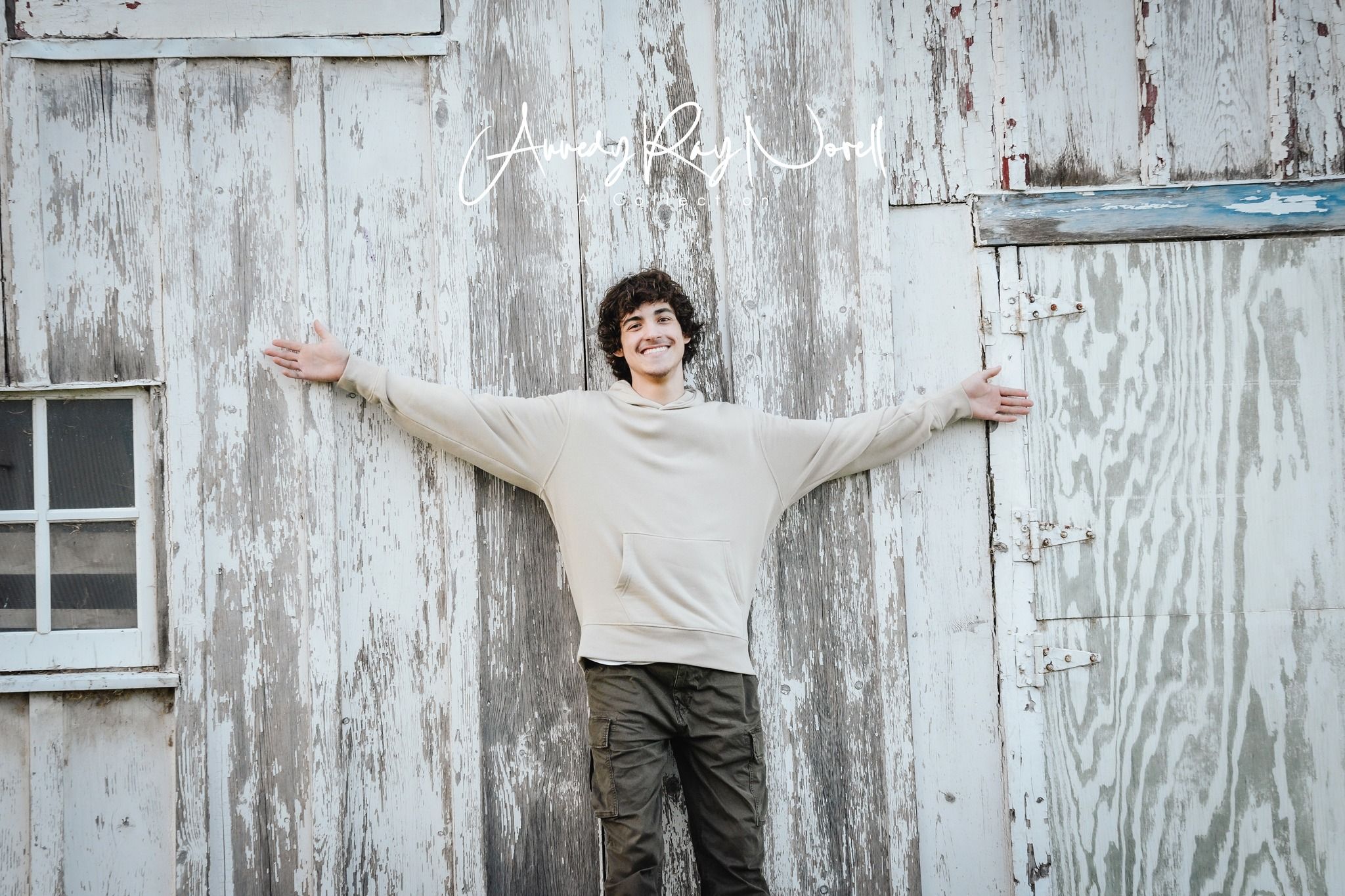 Young man in cream sweater and olive pants stands arms outstretched against rustic white weathered barn wall.