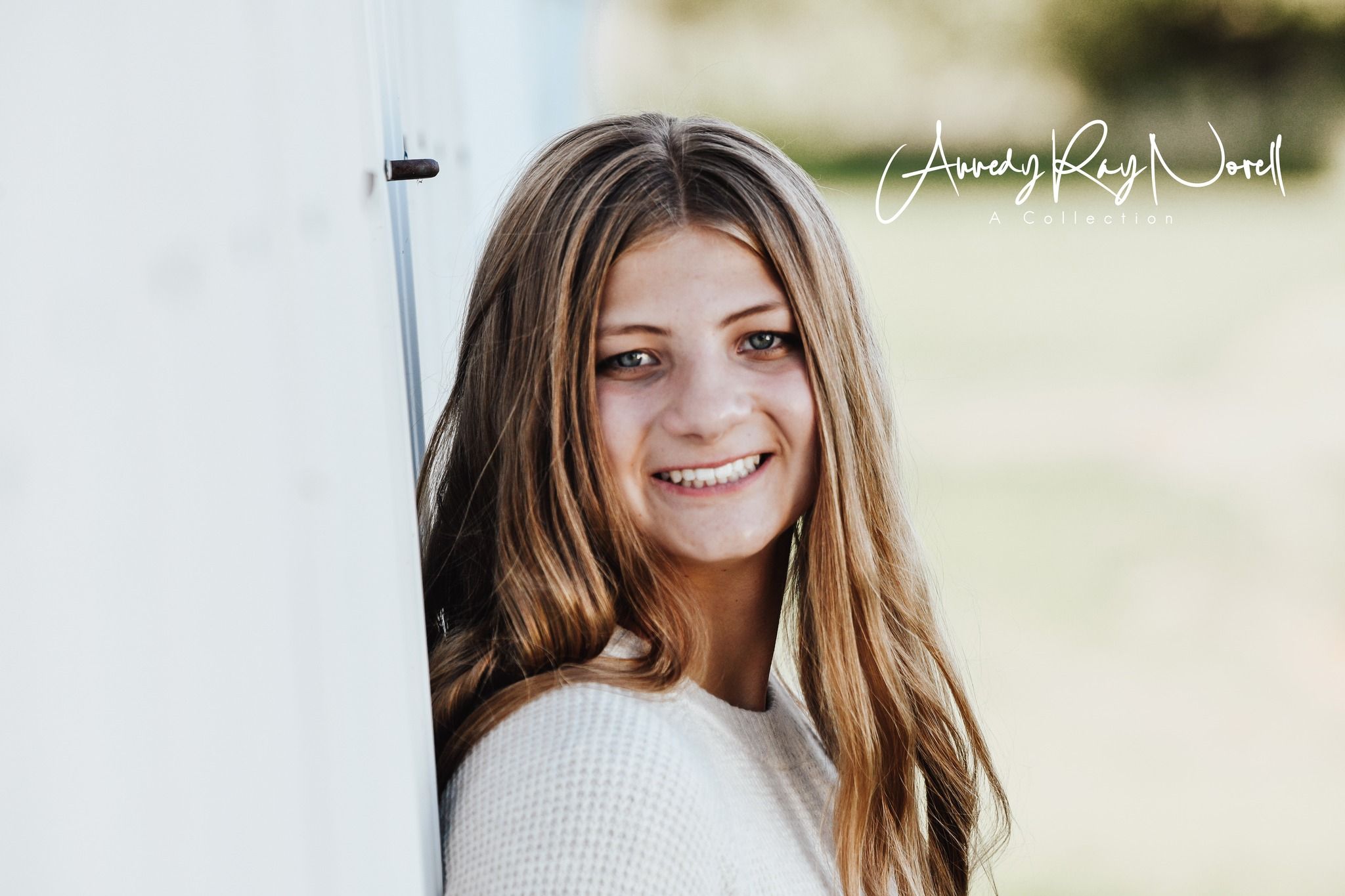 Smiling teenage girl with long brown hair leaning against a white wall in a cream sweater for senior portraits.