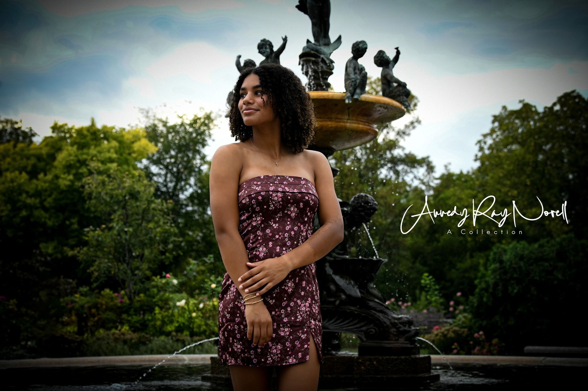 Young woman in floral strapless romper poses before ornate fountain in lush park setting.