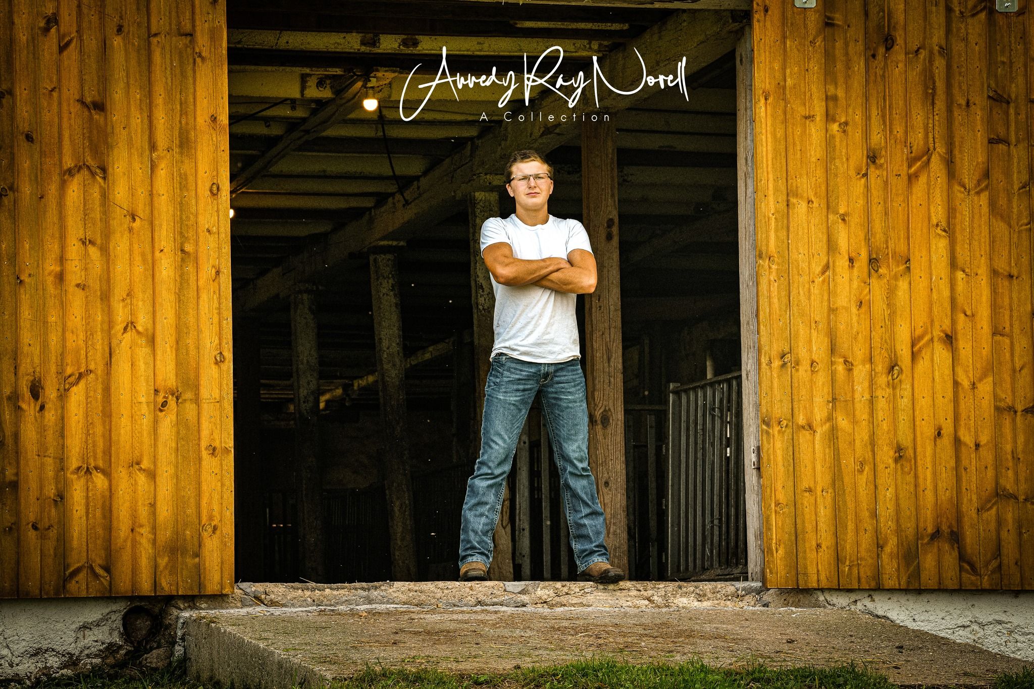Young man in white t-shirt and jeans standing confidently in a rustic barn doorway for senior photos.