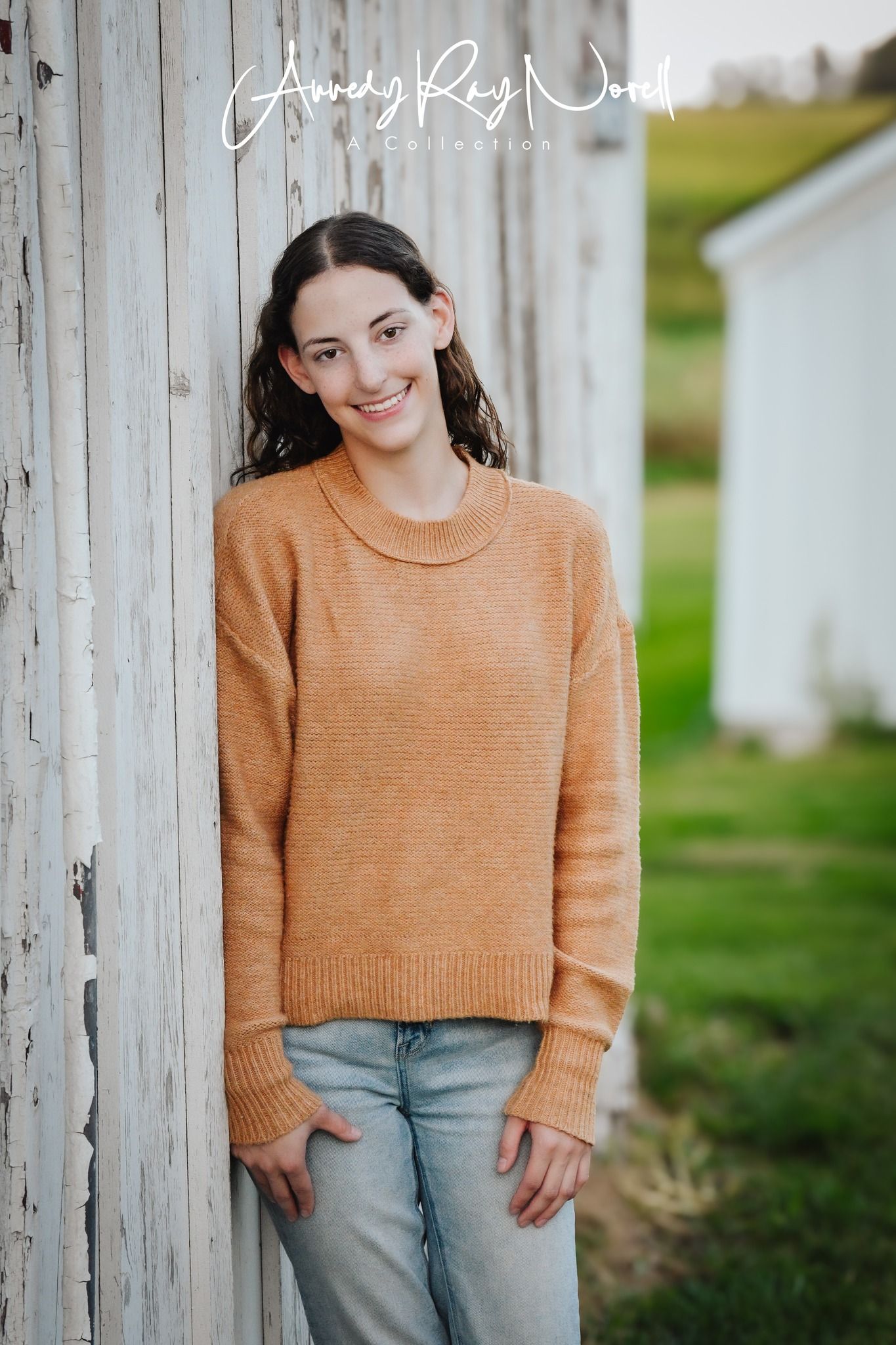 Teen girl in orange sweater and jeans leaning against rustic barn wall.