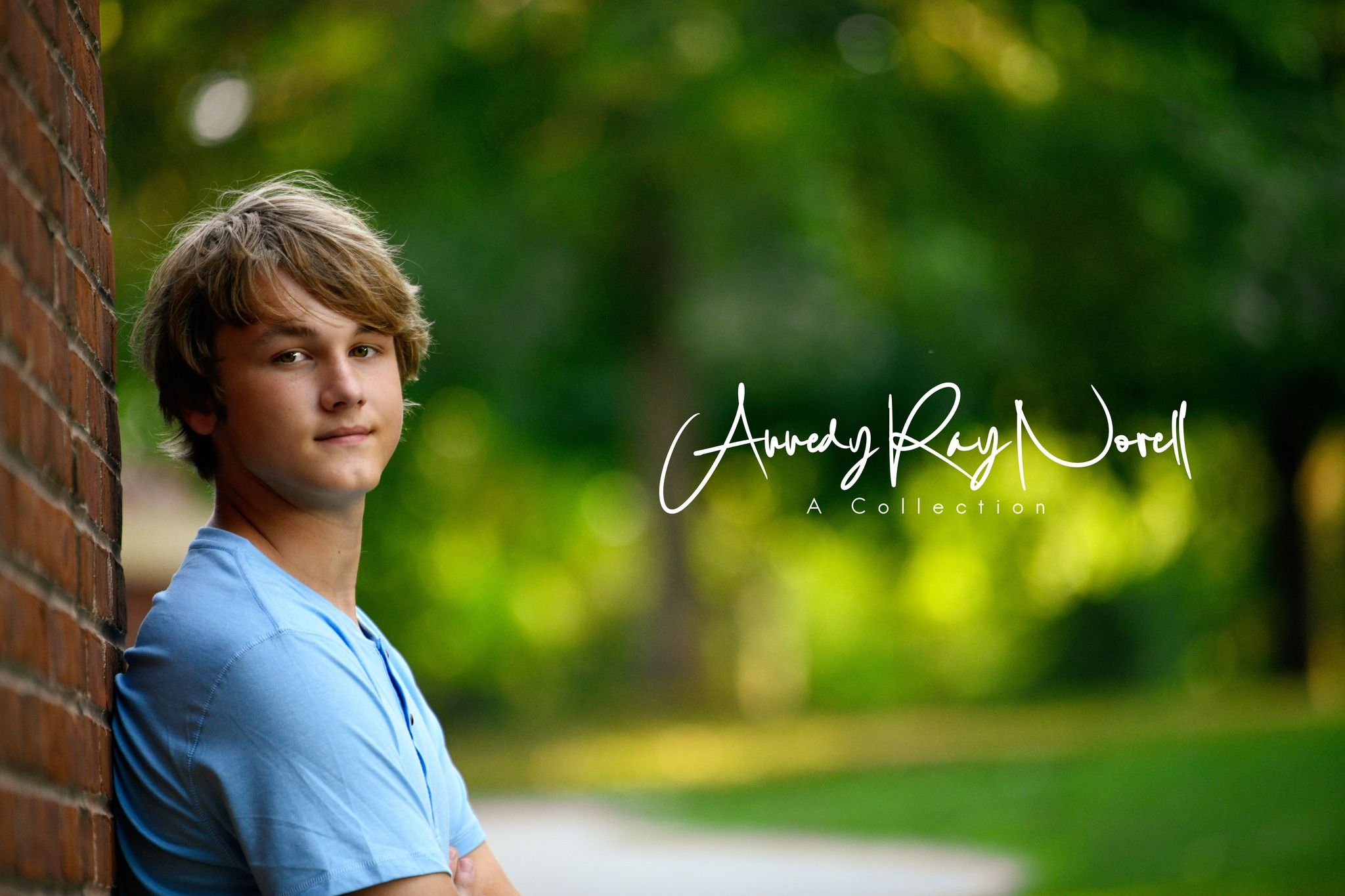 A blonde teenage boy leans against a brick wall with arms crossed in a blue shirt outdoors.