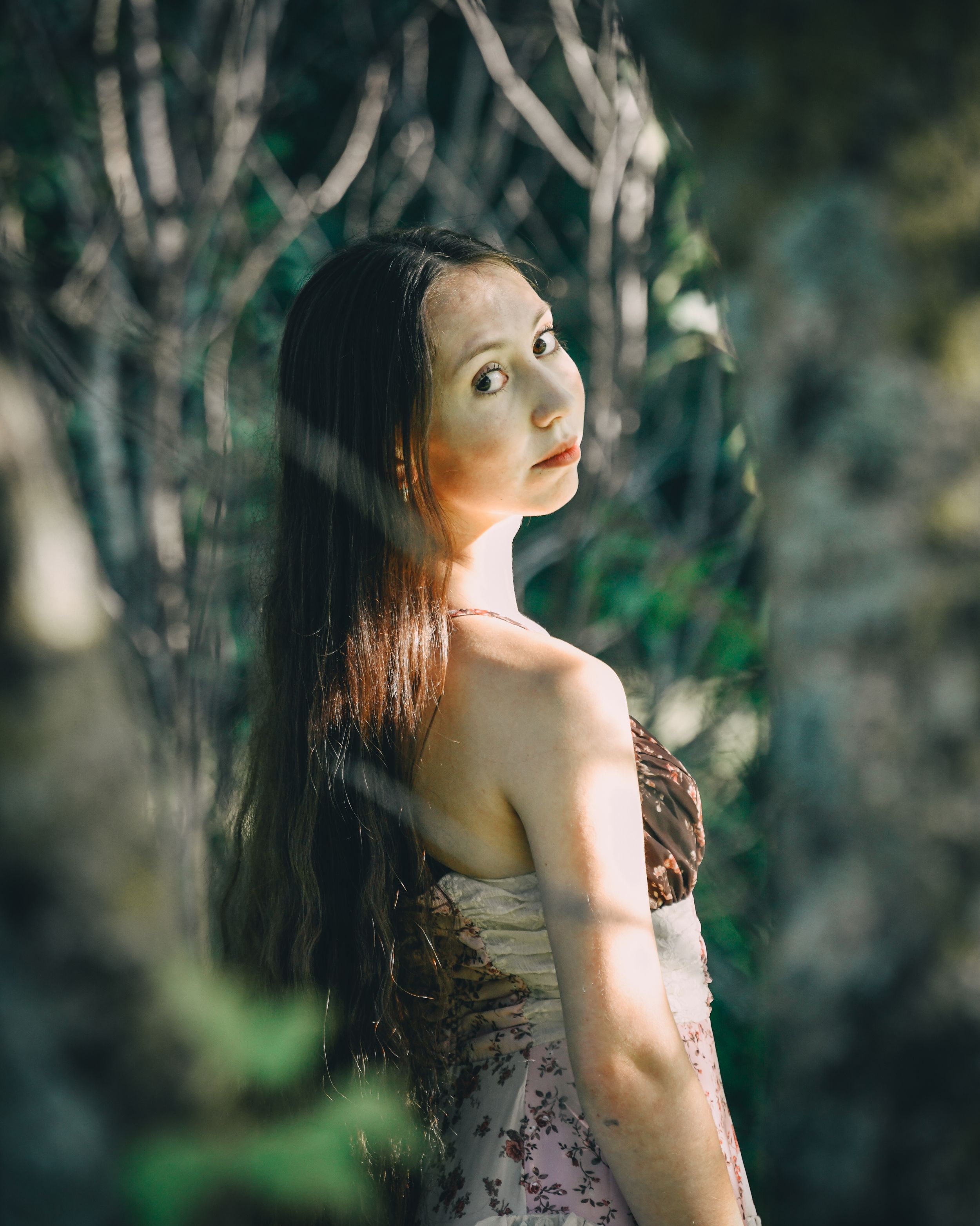 Young woman with long hair in floral dress poses among lush green foliage in soft natural light.