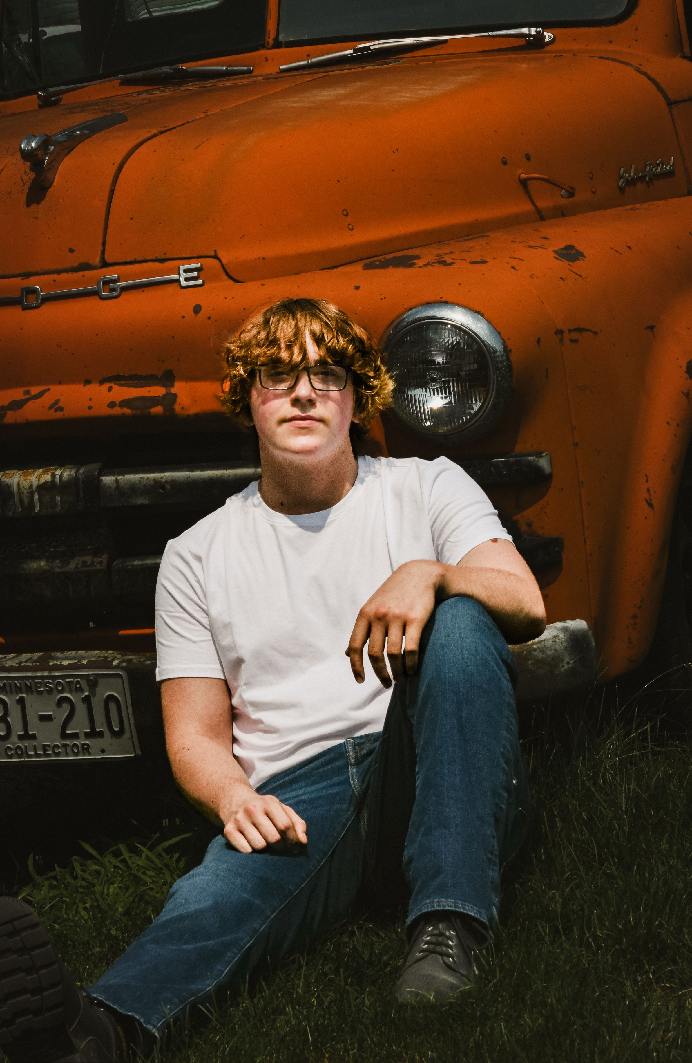 A young man with curly red hair and glasses sits on grass leaning against a vintage orange Dodge truck.