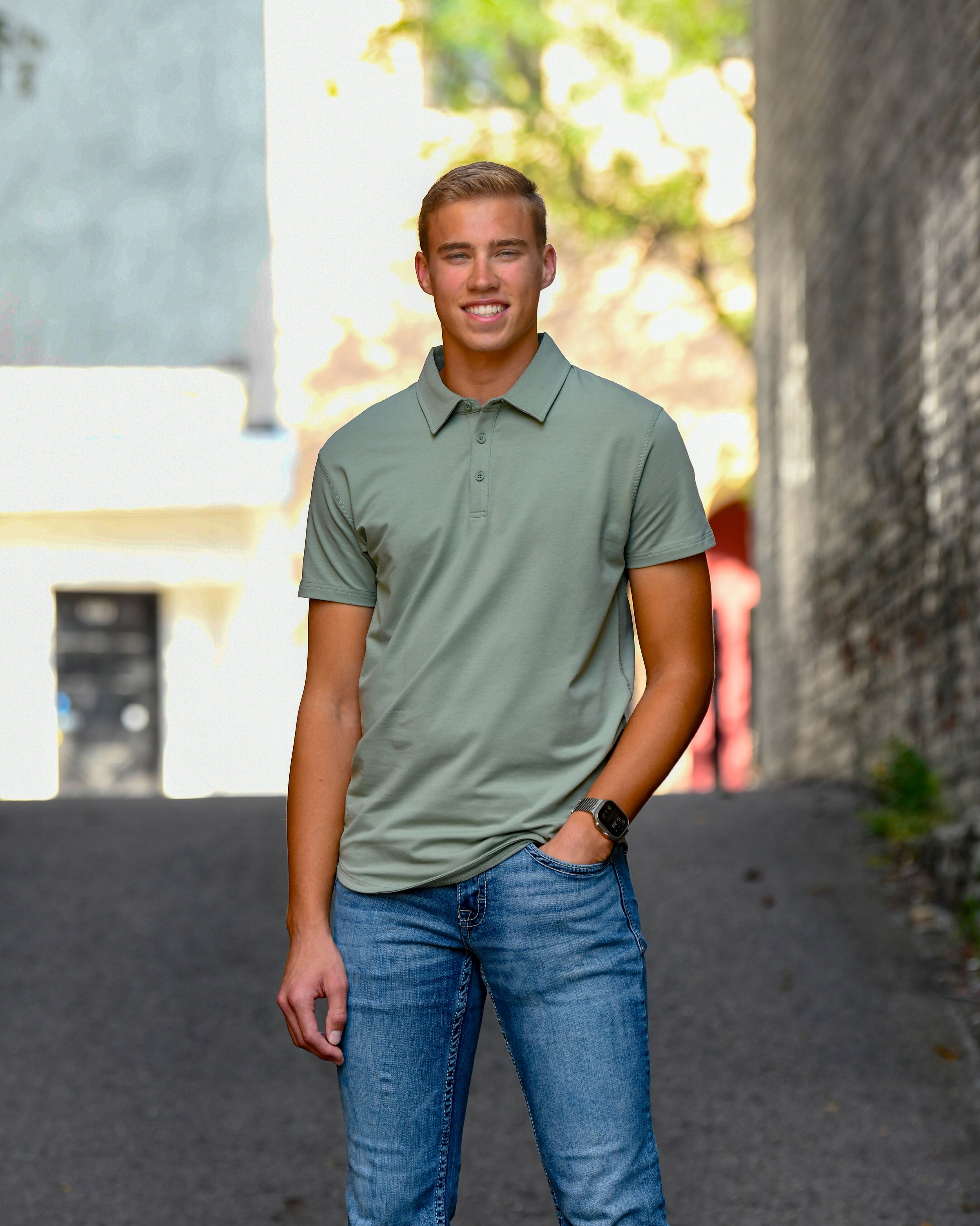 Young man smiling in sage green polo shirt and jeans standing in urban alleyway.